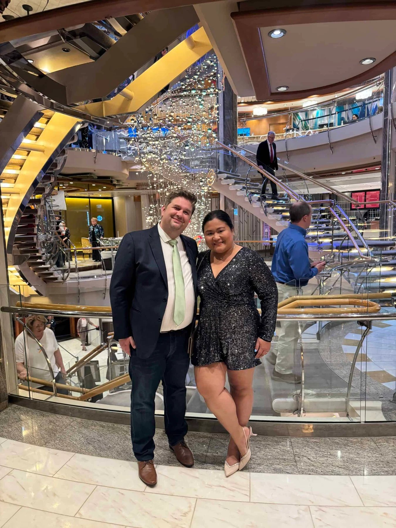 A couple in smart evening wear pose smiling in a grand cruise ship atrium with a sweeping staircase and chandelier.