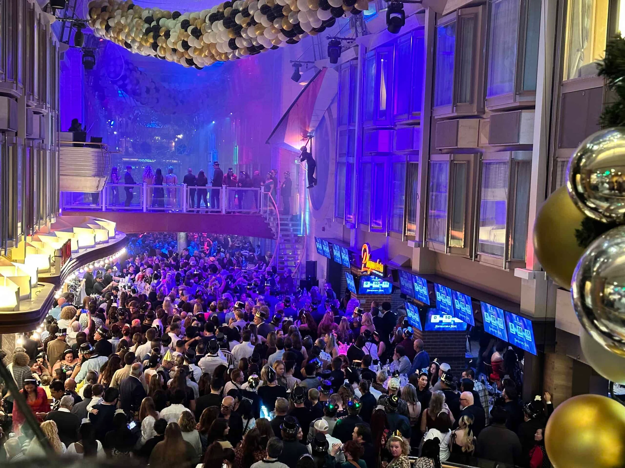 A large crowd fills a multi-deck indoor atrium decorated with black and gold balloons under purple lights at a New Year party.
