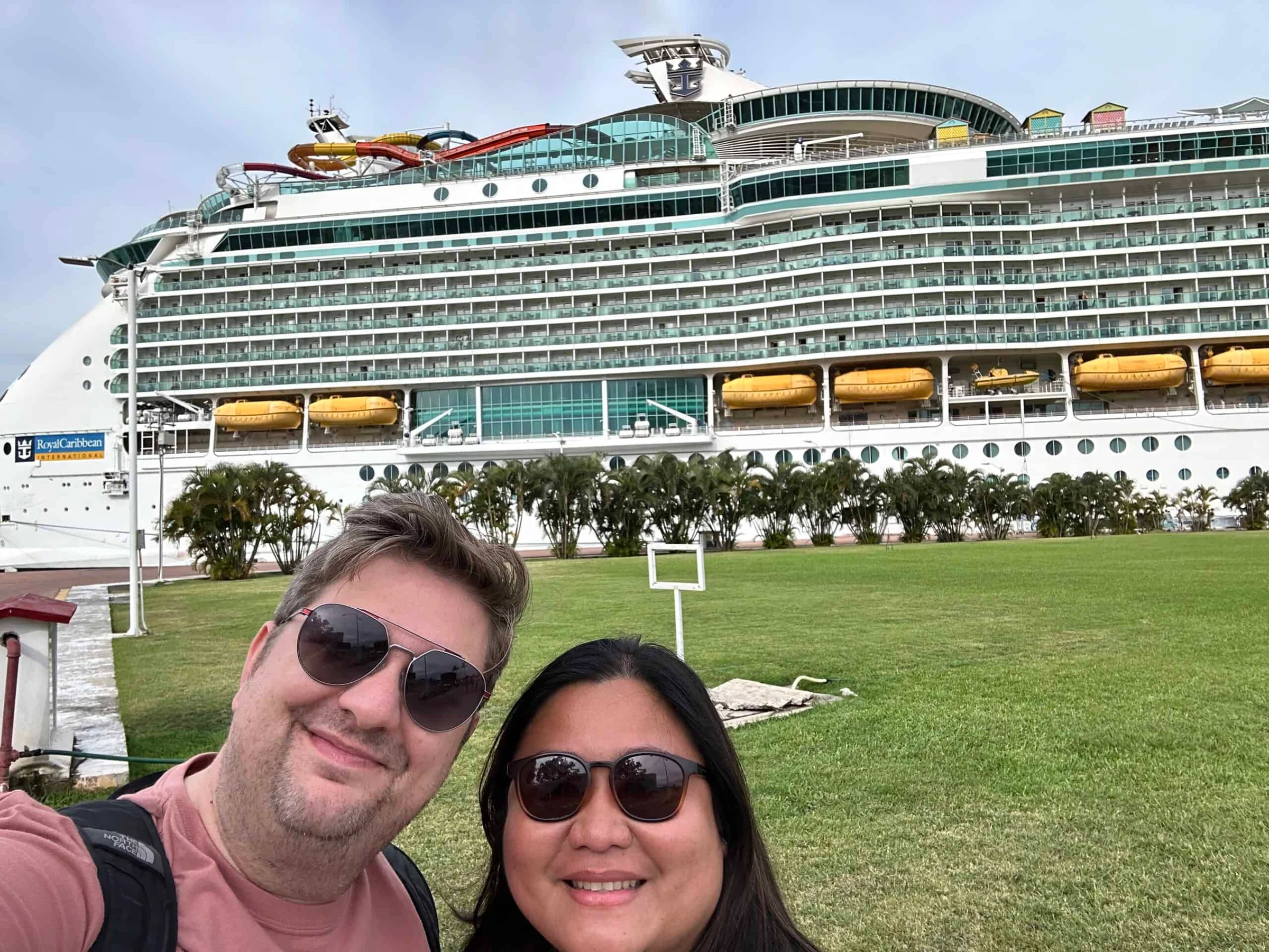 A couple smile for a selfie in front of a large Royal Caribbean cruise ship docked at a port.