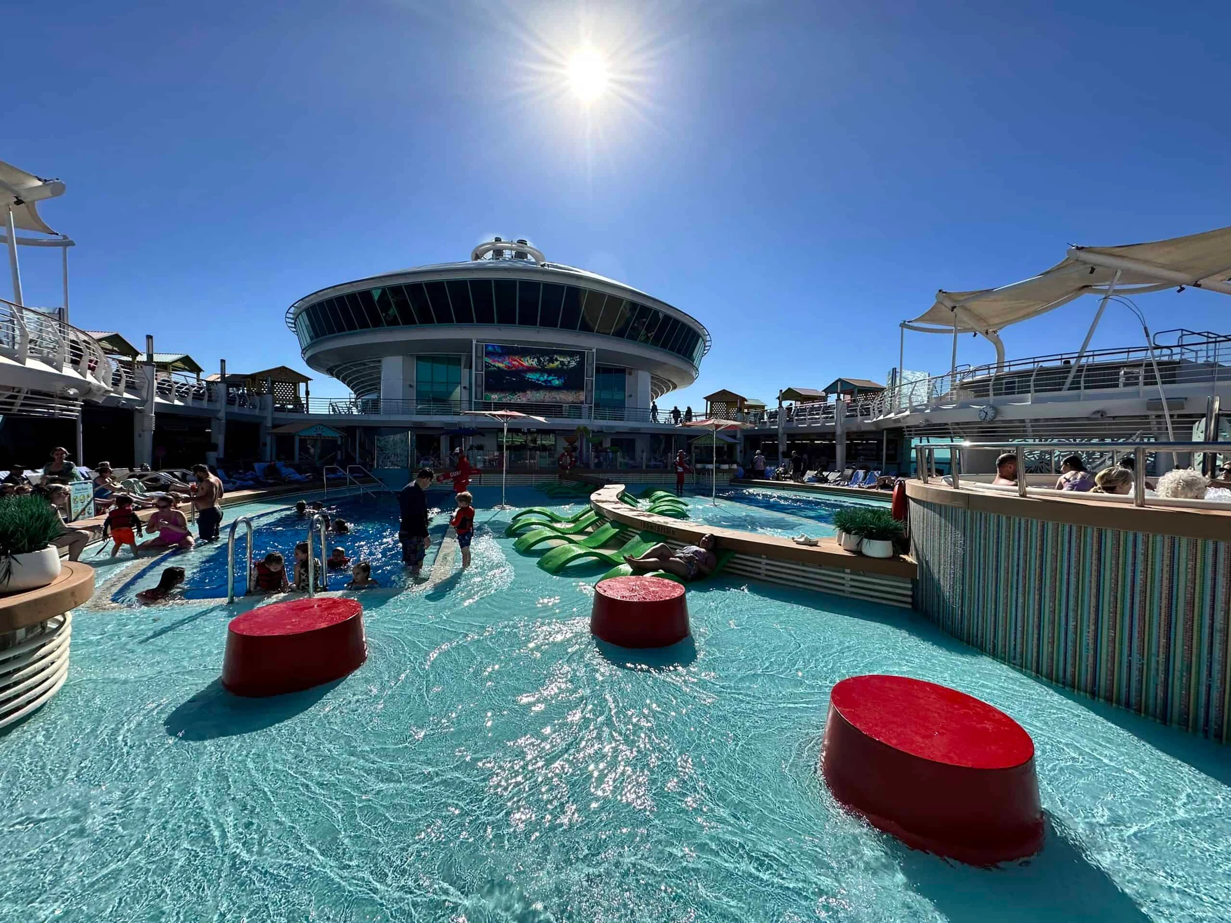 Sunlit cruise ship pool deck with red stepping stones in the water, green loungers and a circular observation deck behind.