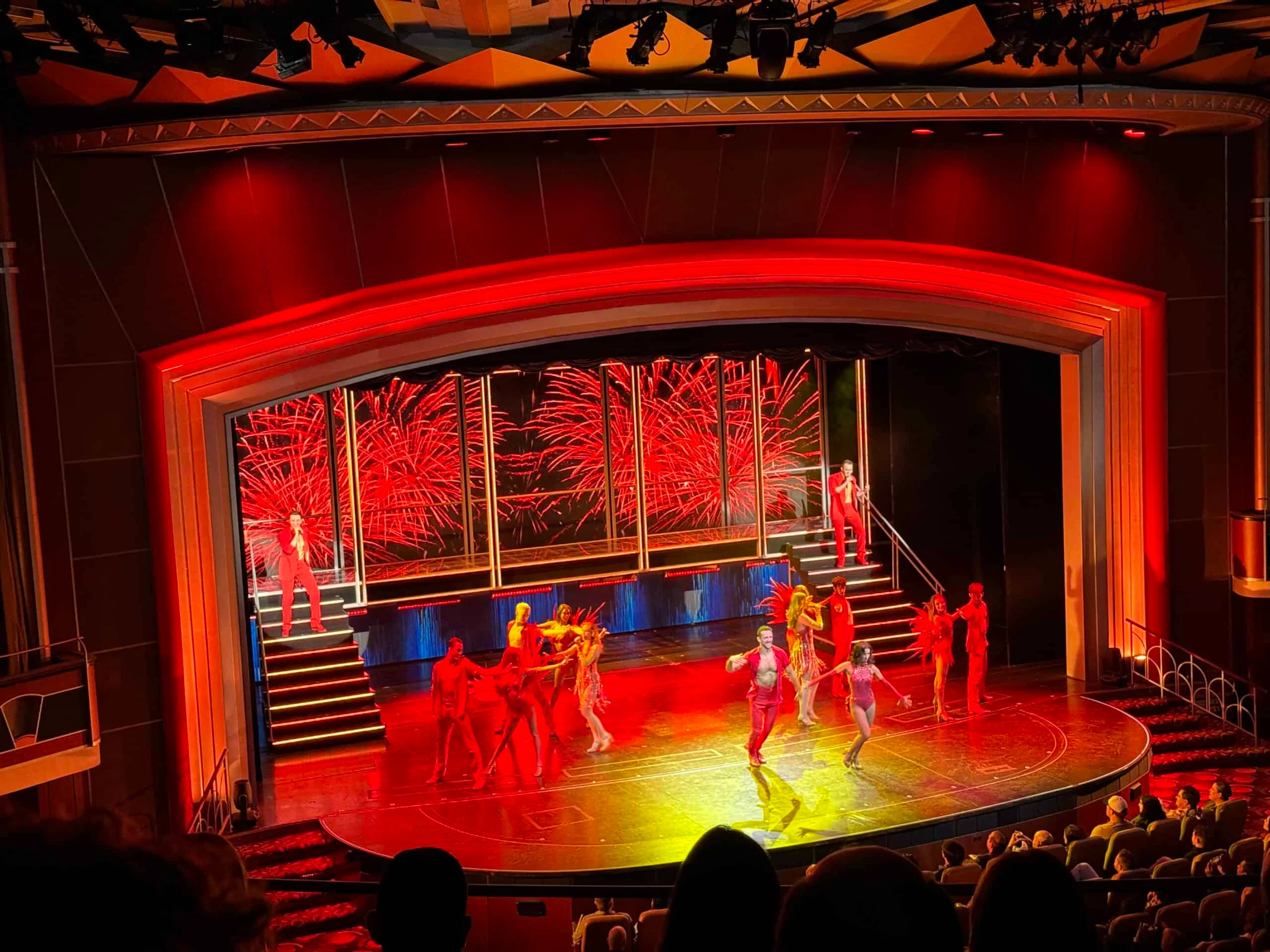 Performers in red costumes dance on a theatre stage lit in deep red, with a fireworks backdrop behind them.