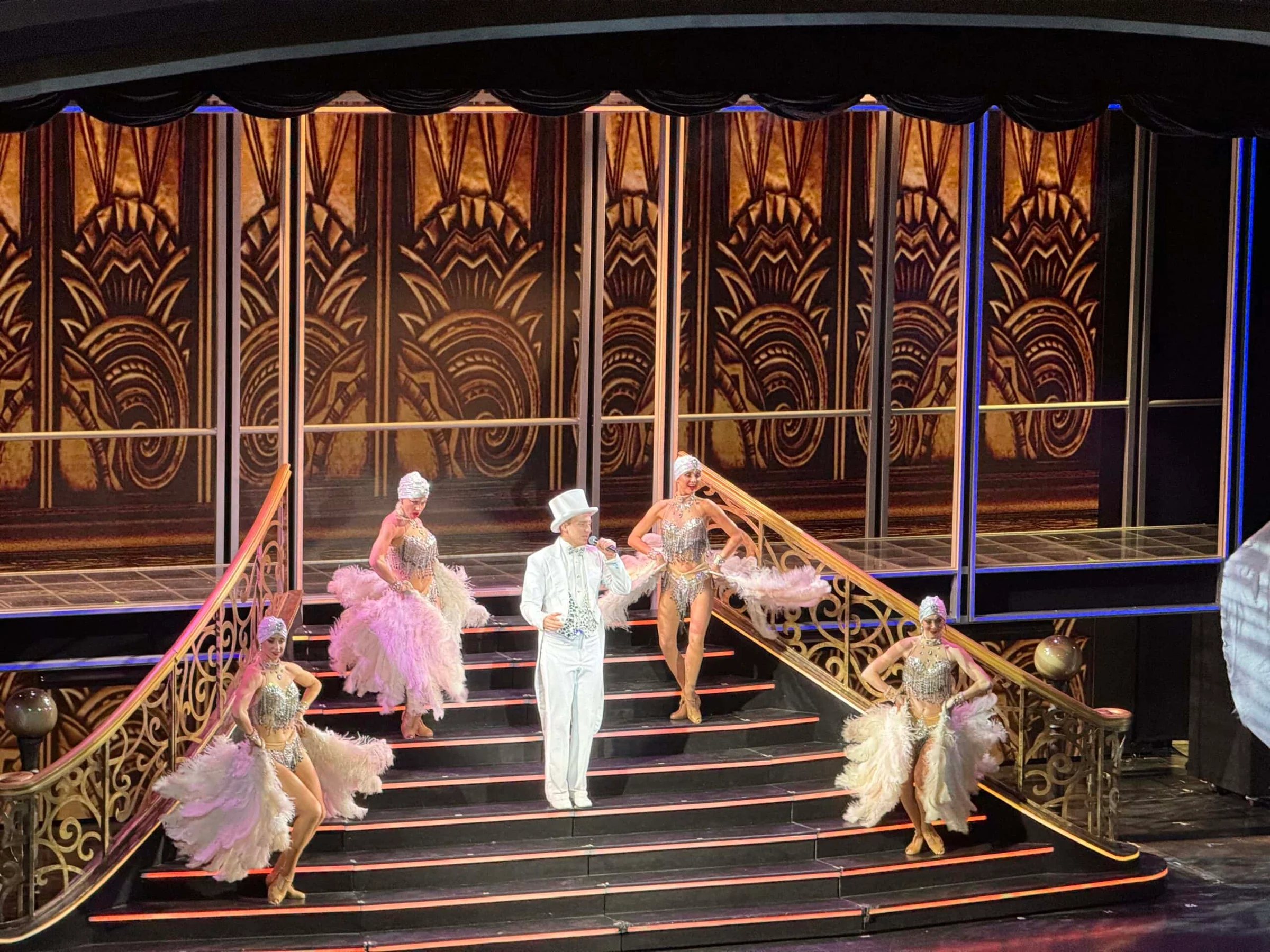 A performer in a white tuxedo and top hat sings on a grand staircase stage flanked by four showgirls in feathered costumes.