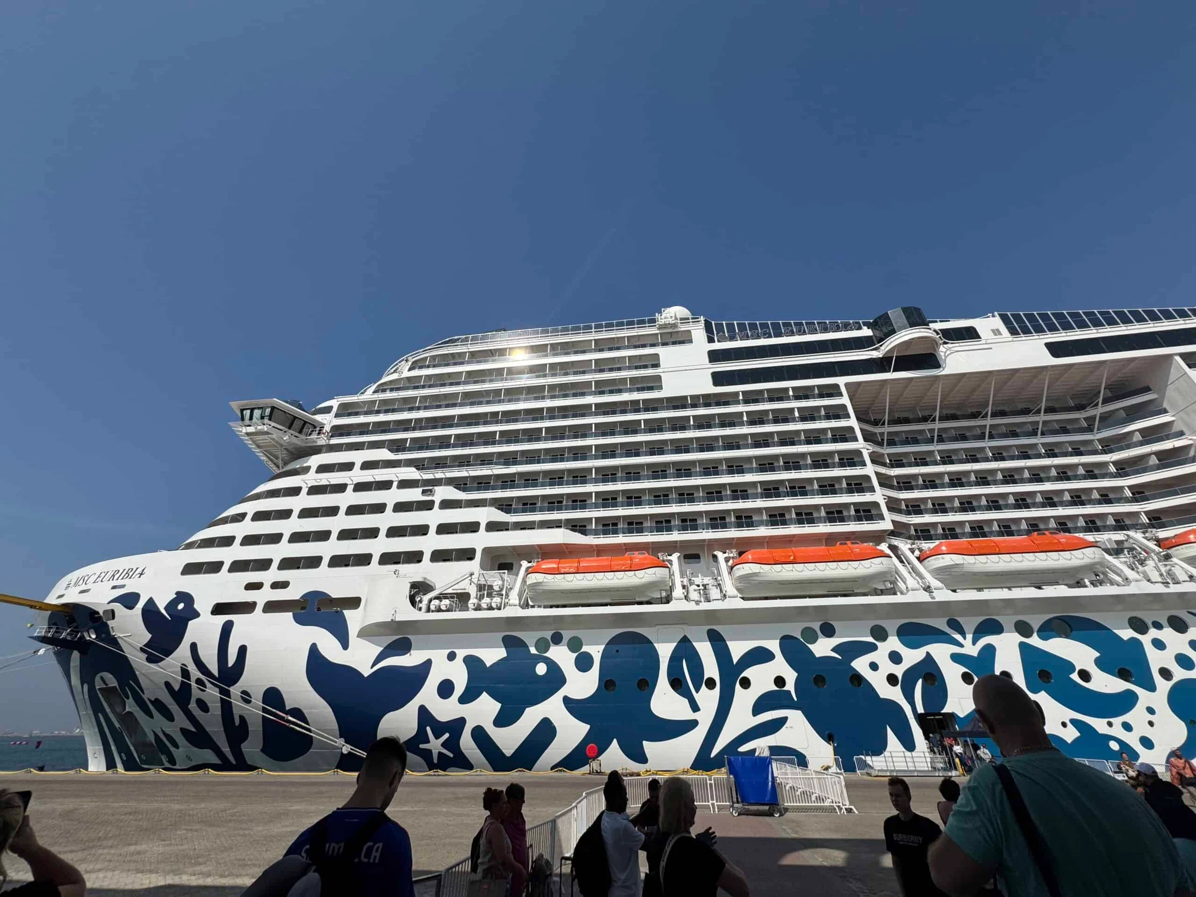 The MSC Euribia cruise ship docked at a port, its white hull decorated with bold blue marine patterns, with passengers gathered on the quayside below.
