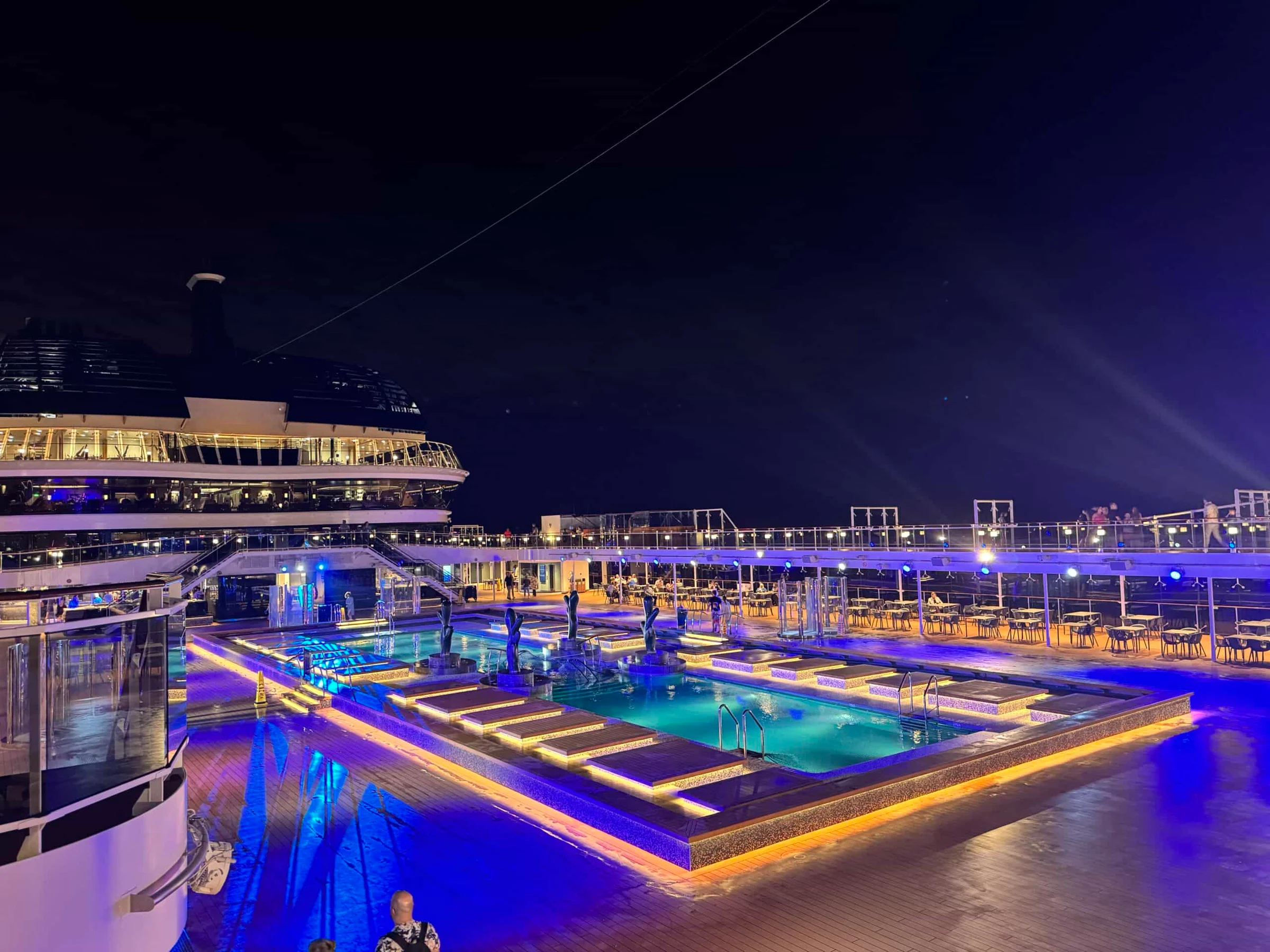 A cruise ship pool deck lit with blue and amber lights at night, with sun loungers, seating areas and upper decks visible.