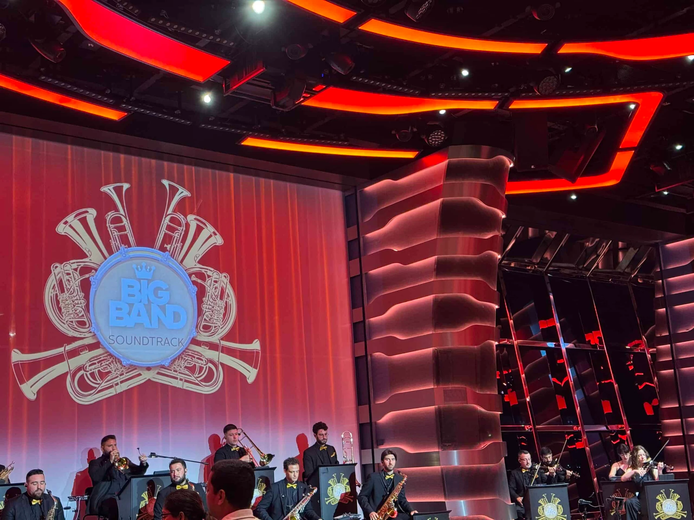 Musicians in black suits with yellow bow ties perform on a stage beneath a Big Band Soundtrack logo on a red screen.