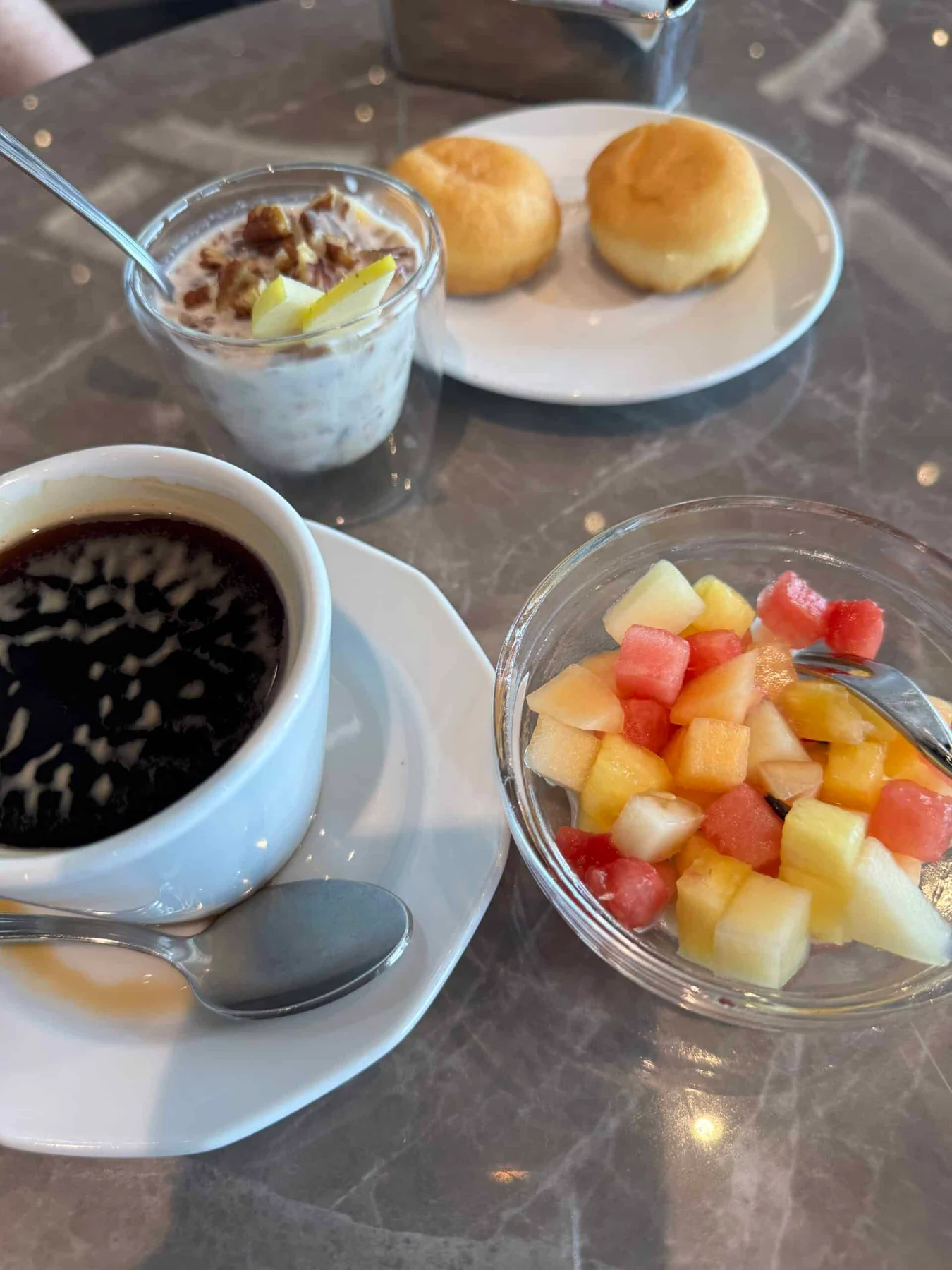 A marble table set with black coffee, a fruit salad, a creamy dessert with nuts and a plate of bread rolls.