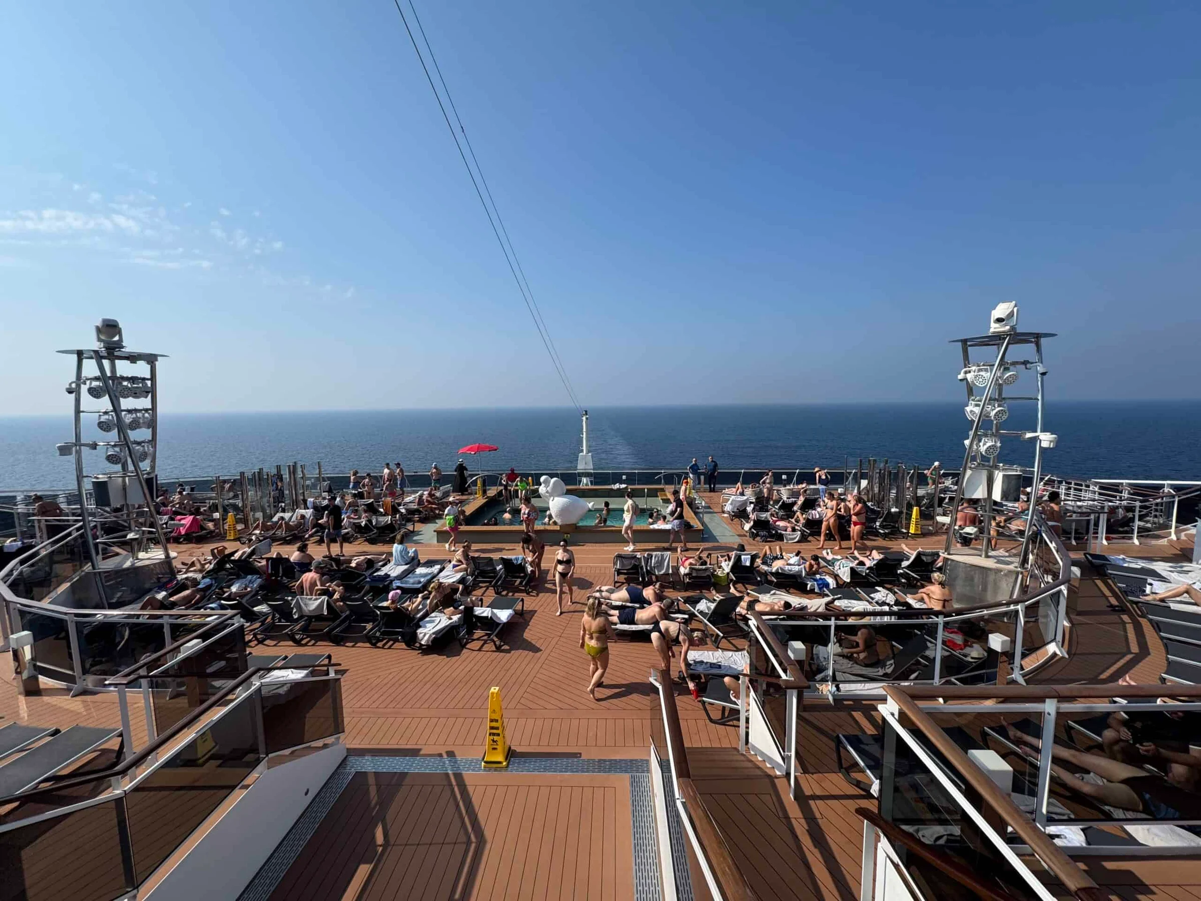 Passengers sunbathing on packed loungers across the upper deck of a cruise ship sailing on calm open sea.