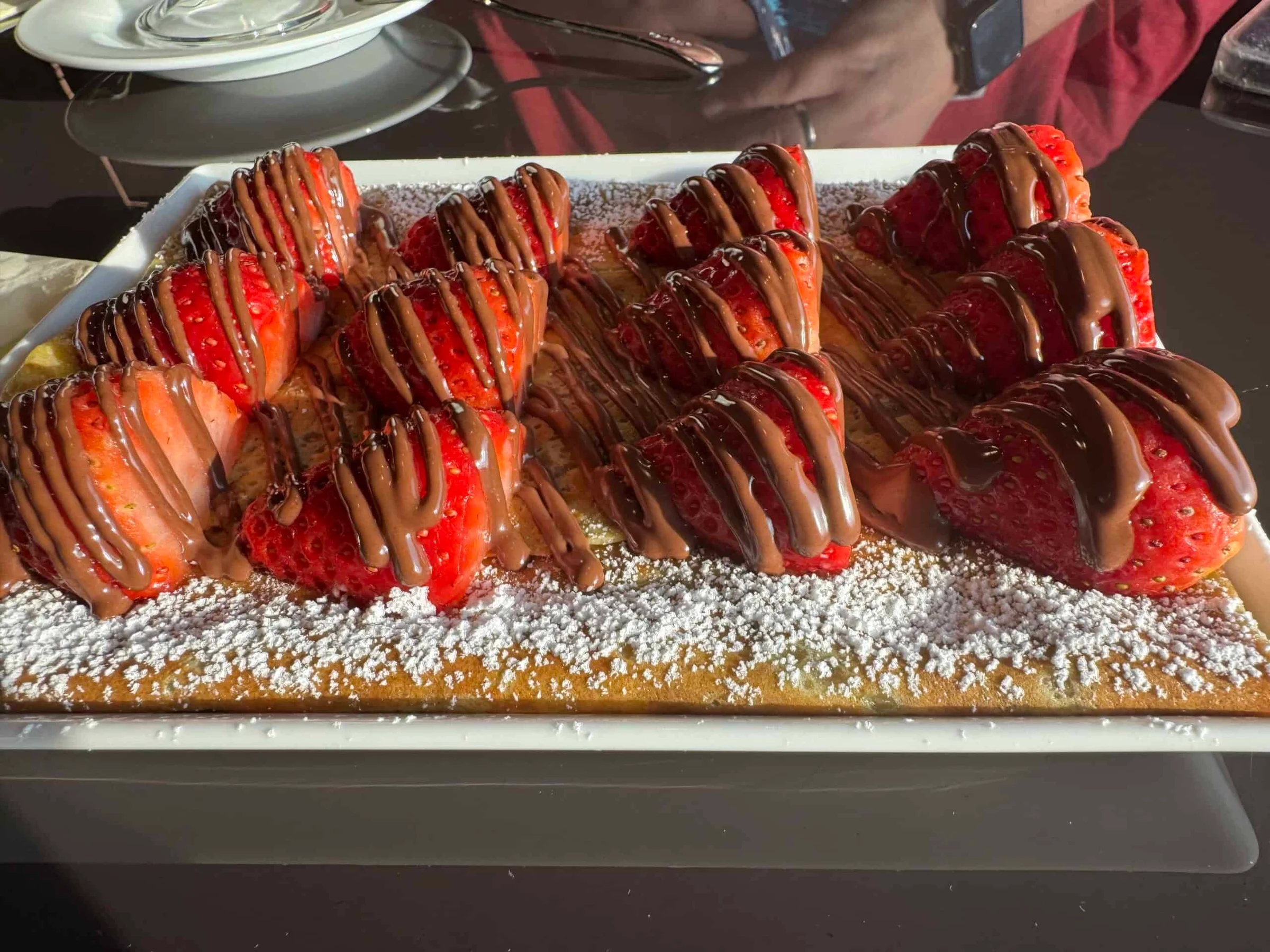 Sliced strawberries drizzled with chocolate sauce arranged on a sponge base dusted with icing sugar on a white rectangular plate.