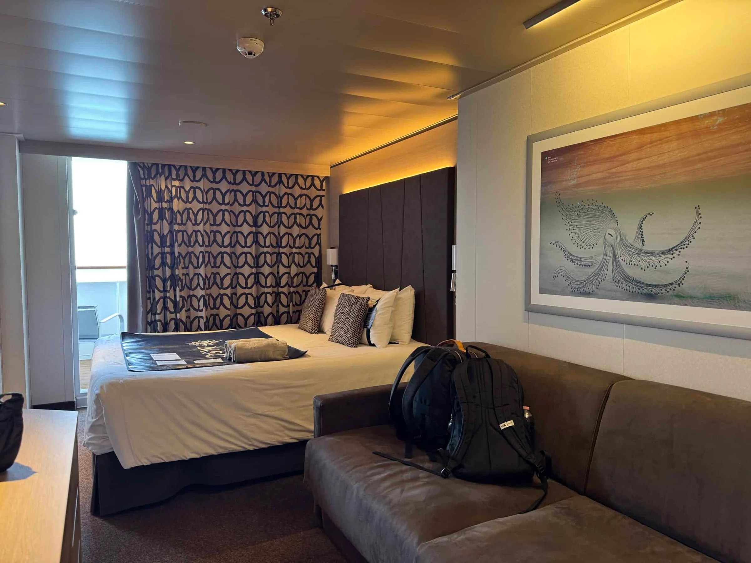 A cruise ship cabin with a double bed, patterned curtains, a sofa with a backpack on it and an octopus artwork on the wall.