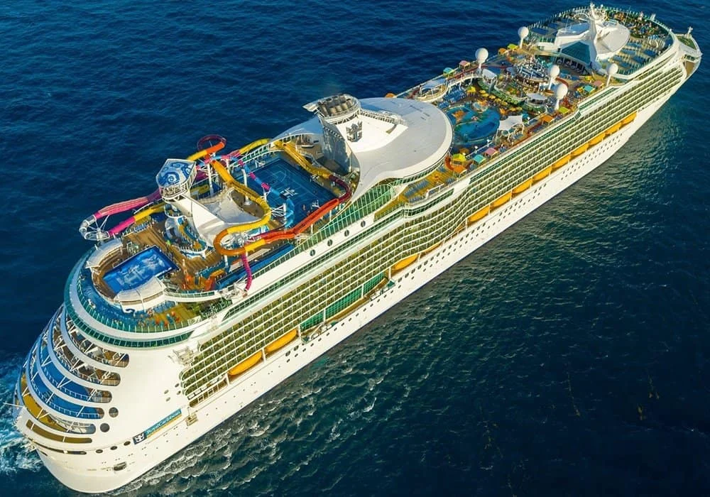 Aerial view of a large white cruise ship with water slides and deck amenities sailing on dark blue open sea.