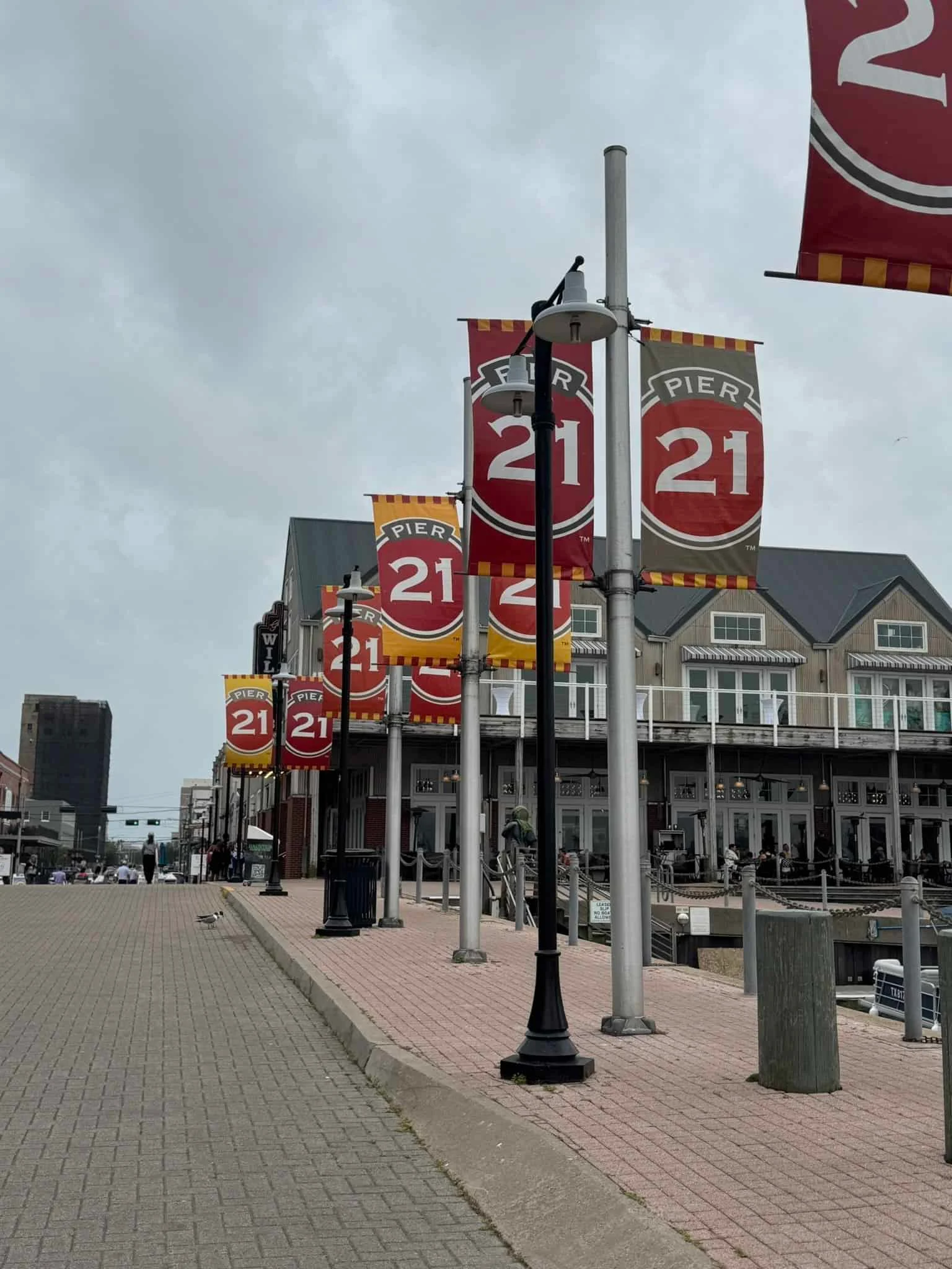 A brick waterfront promenade lined with lamp posts bearing red and yellow Pier 21 banners, with a large building behind.