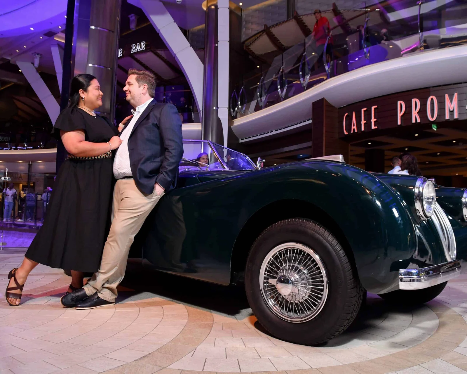 A couple lean against a dark green vintage convertible inside a cruise ship atrium near a Cafe Promenade sign.