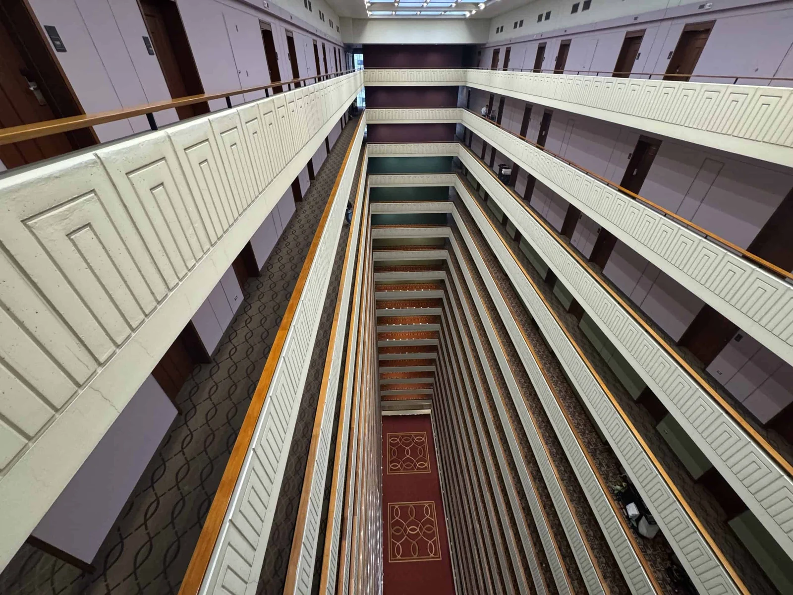 A downward view into a multi-storey hotel atrium with white balconies, wooden railings, and a red patterned carpet far below.