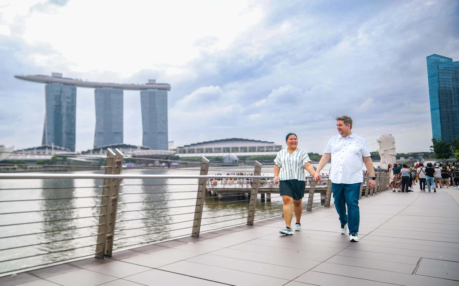A couple walk hand in hand along a waterfront promenade with Marina Bay Sands visible behind them.