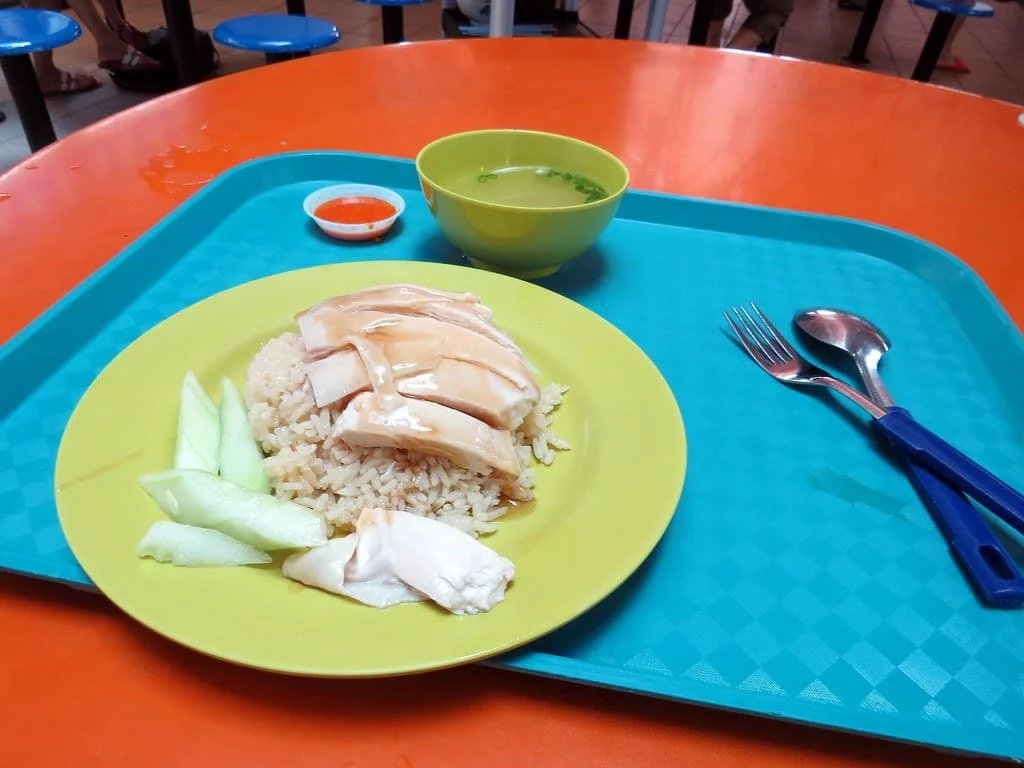 A plate of Hainanese chicken rice with cucumber slices, a small bowl of soup and chilli sauce on a blue tray.