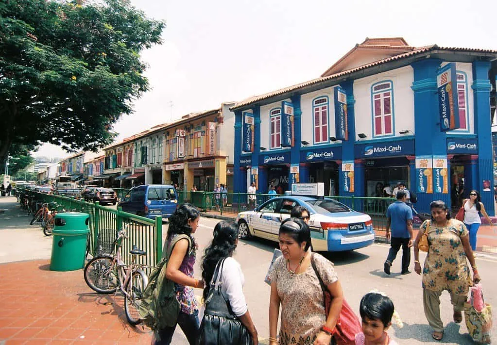 Pedestrians walk along a busy street lined with colourful shophouses, one painted bright blue with Maxi-Cash signs.