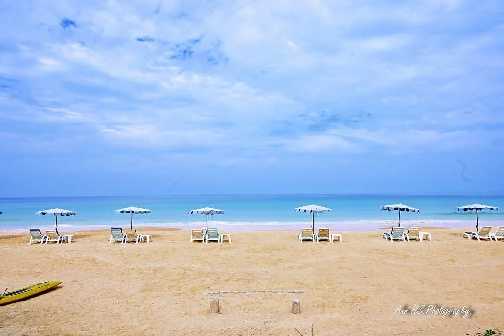 Rows of white sun loungers and striped parasols on a sandy beach facing calm turquoise water under a cloudy blue sky.