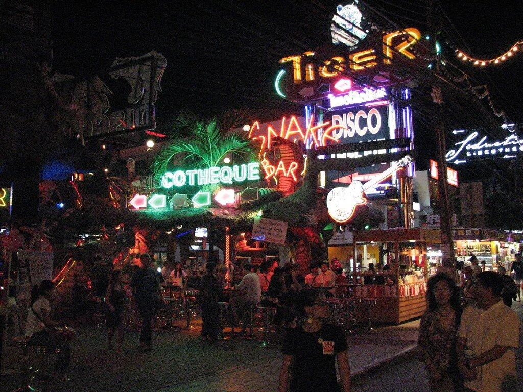 Busy night street lined with neon signs for Tiger Disco and Discotheque Bar, with crowds of people walking past lit-up venues.