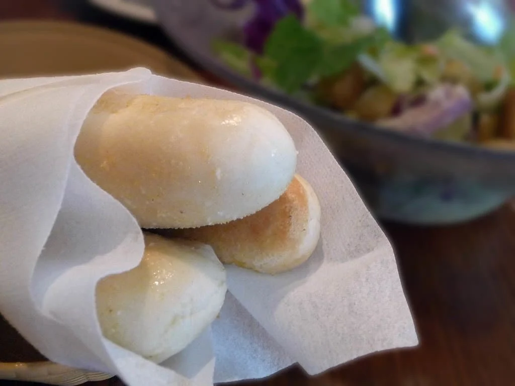 Three small bread rolls served in a paper napkin in a basket, with a salad bowl blurred in the background.