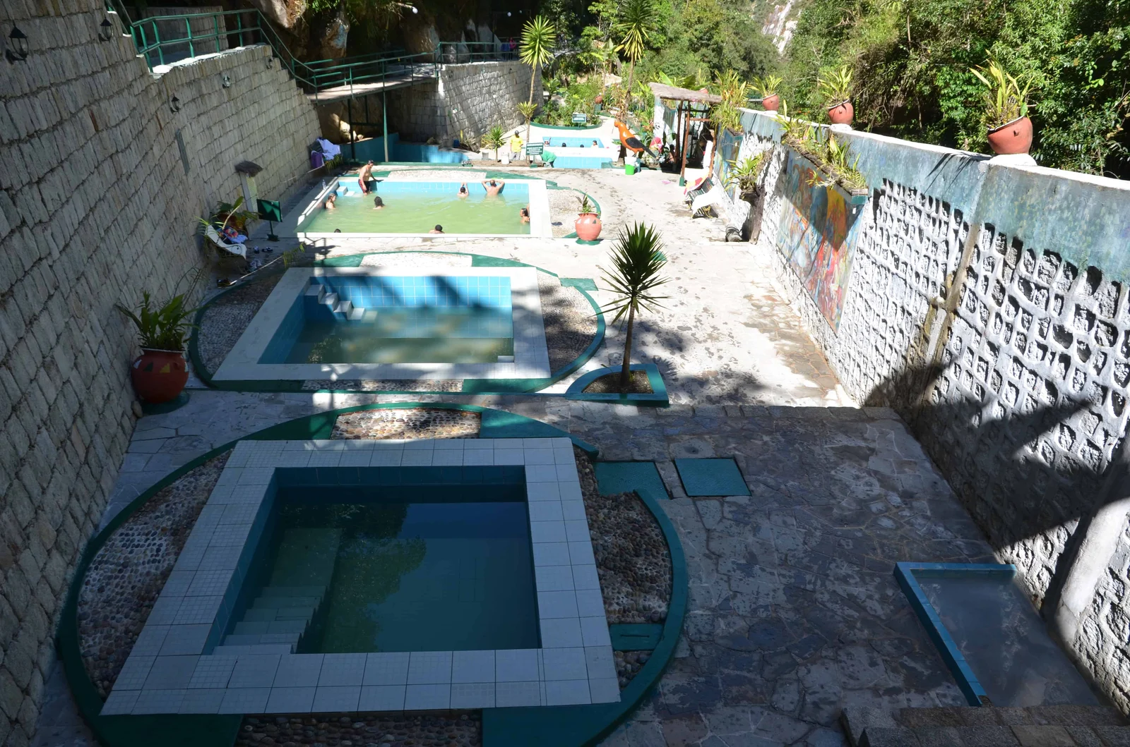 An outdoor tiered pool complex with stone walls, potted plants and lush trees, with people swimming in the upper pool.