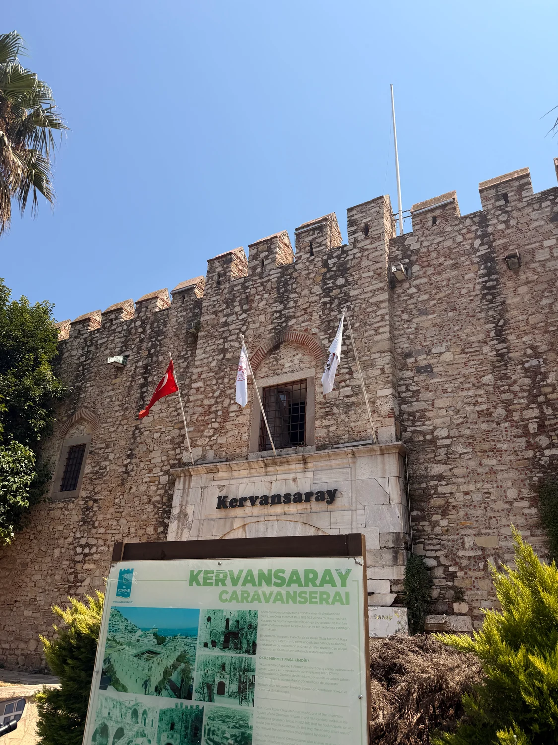 A stone fortress with battlements and three flags flies above a sign reading Kervansaray Caravanserai under a clear blue sky.