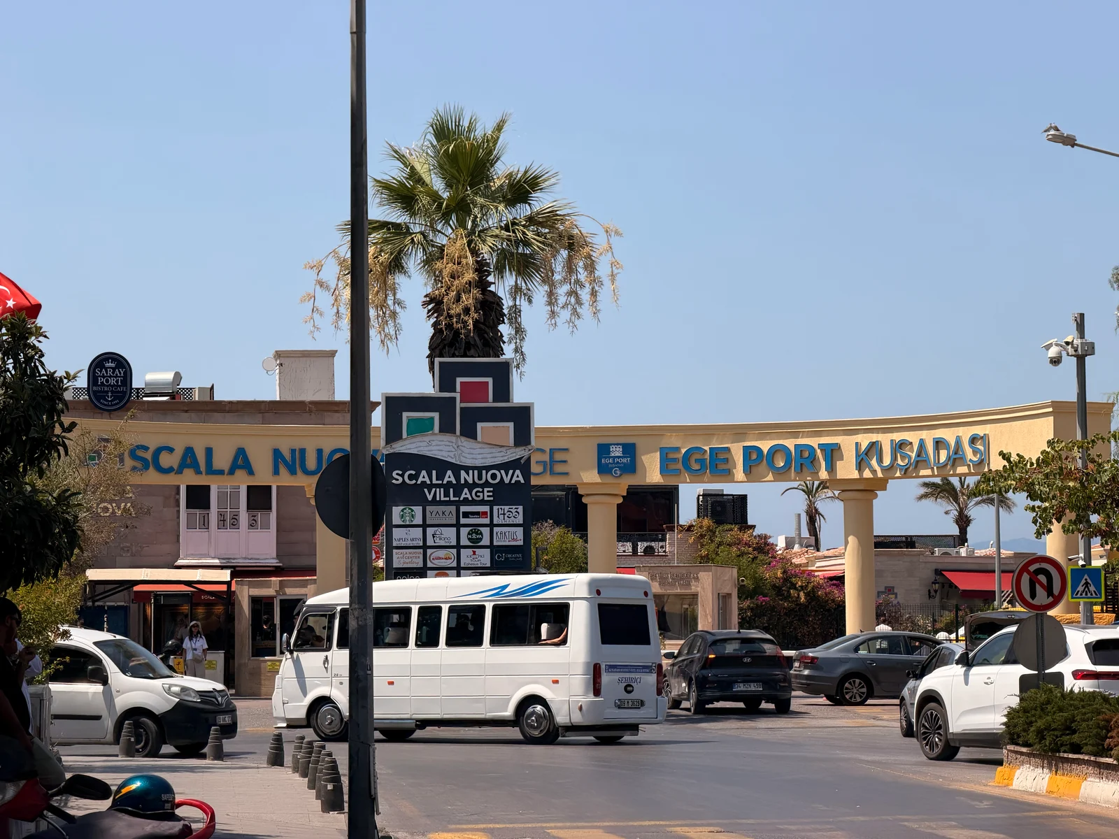 A busy road in front of the Scala Nuova Village shopping complex and Ege Port Kusadasi entrance arch, with cars and a minibus passing by.