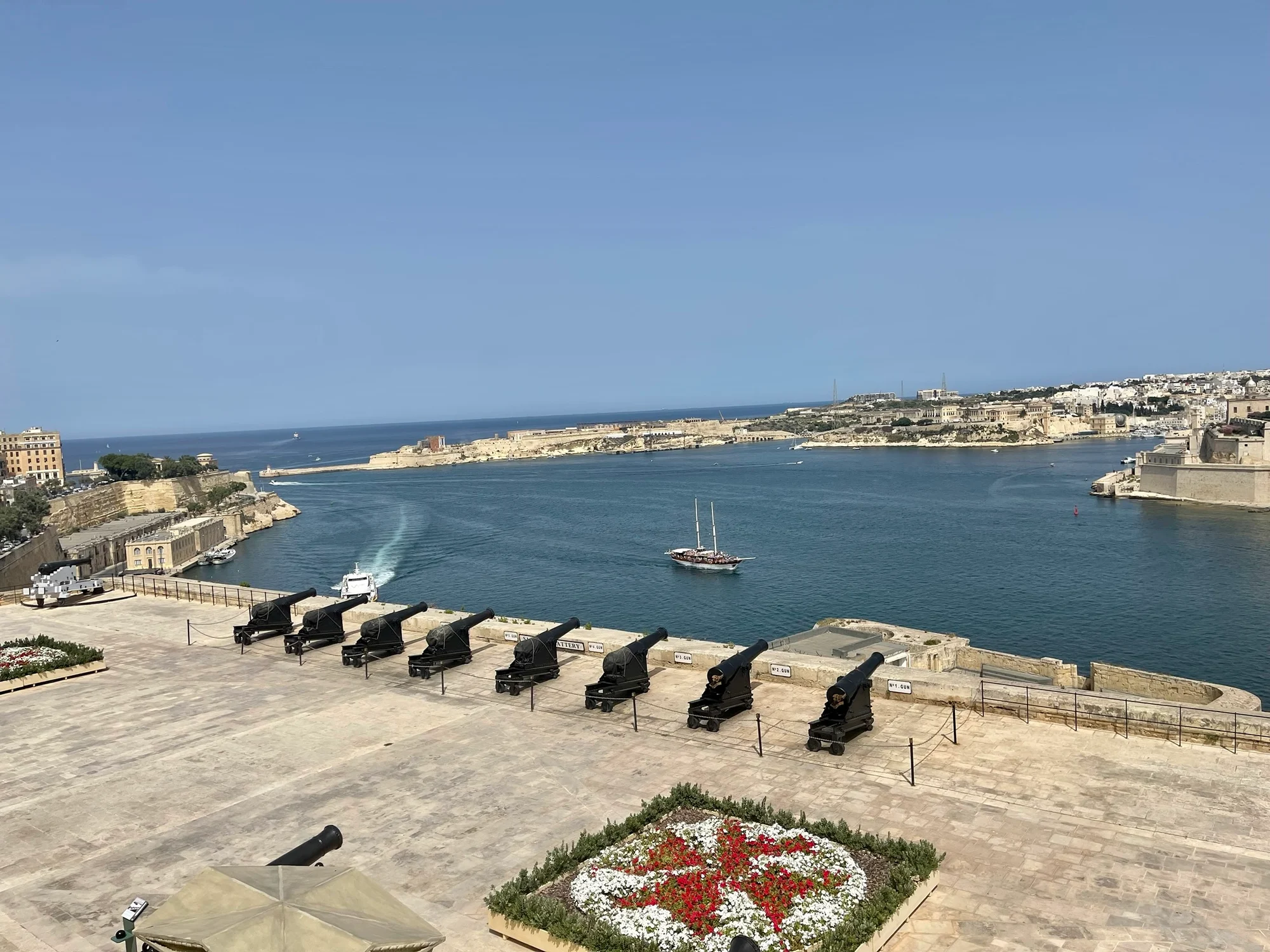 A row of black cannons lines a stone terrace overlooking a broad harbour with boats and a coastal city beyond.