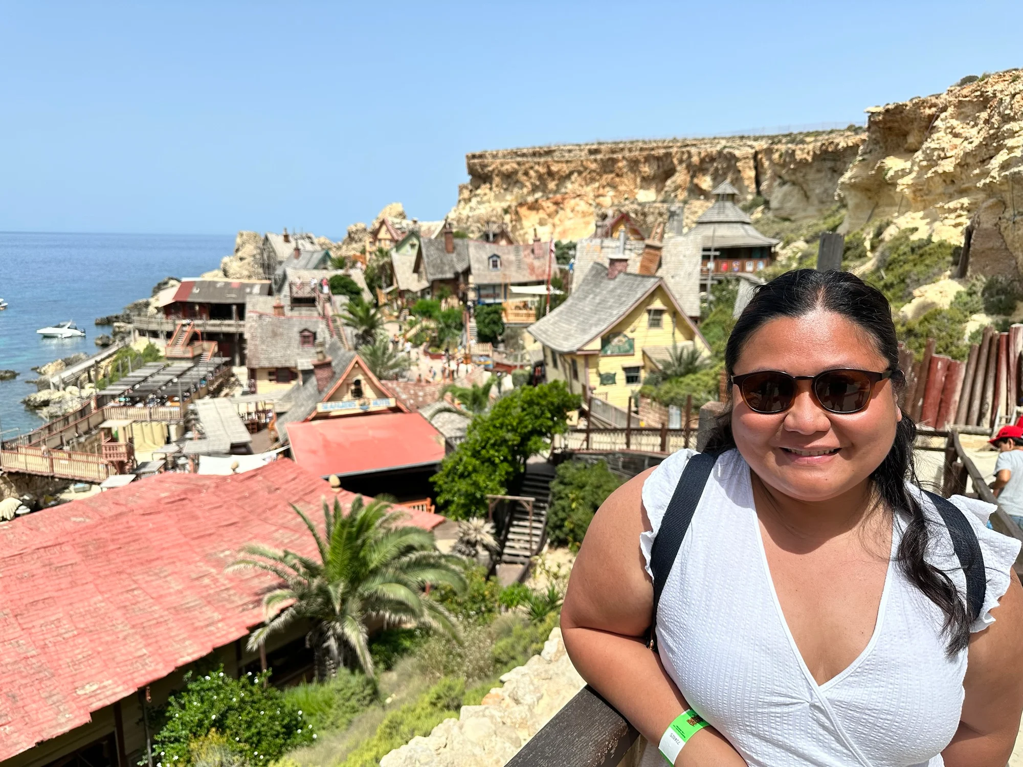 A woman in sunglasses and a white top leans on a railing overlooking a colourful village of wooden buildings beside the sea.