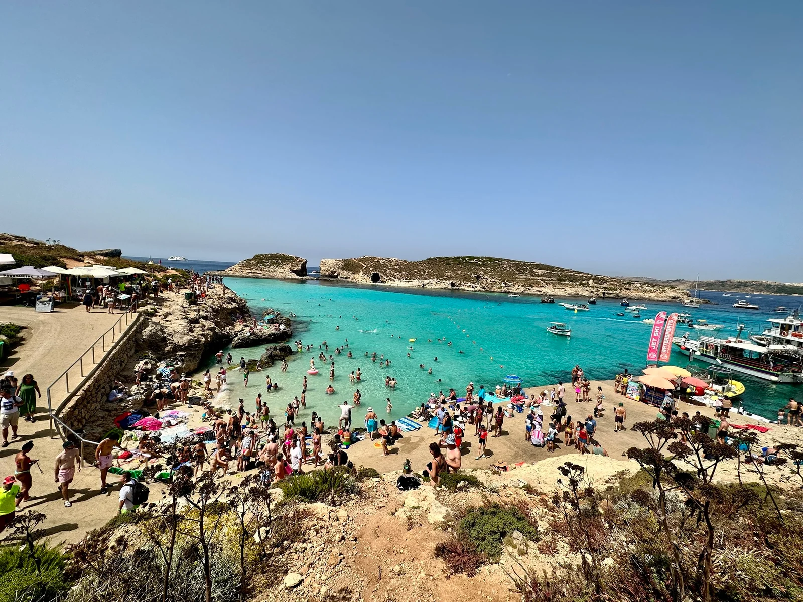 A crowded turquoise bay with swimmers and boats, surrounded by rocky cliffs and a small island under a clear blue sky.