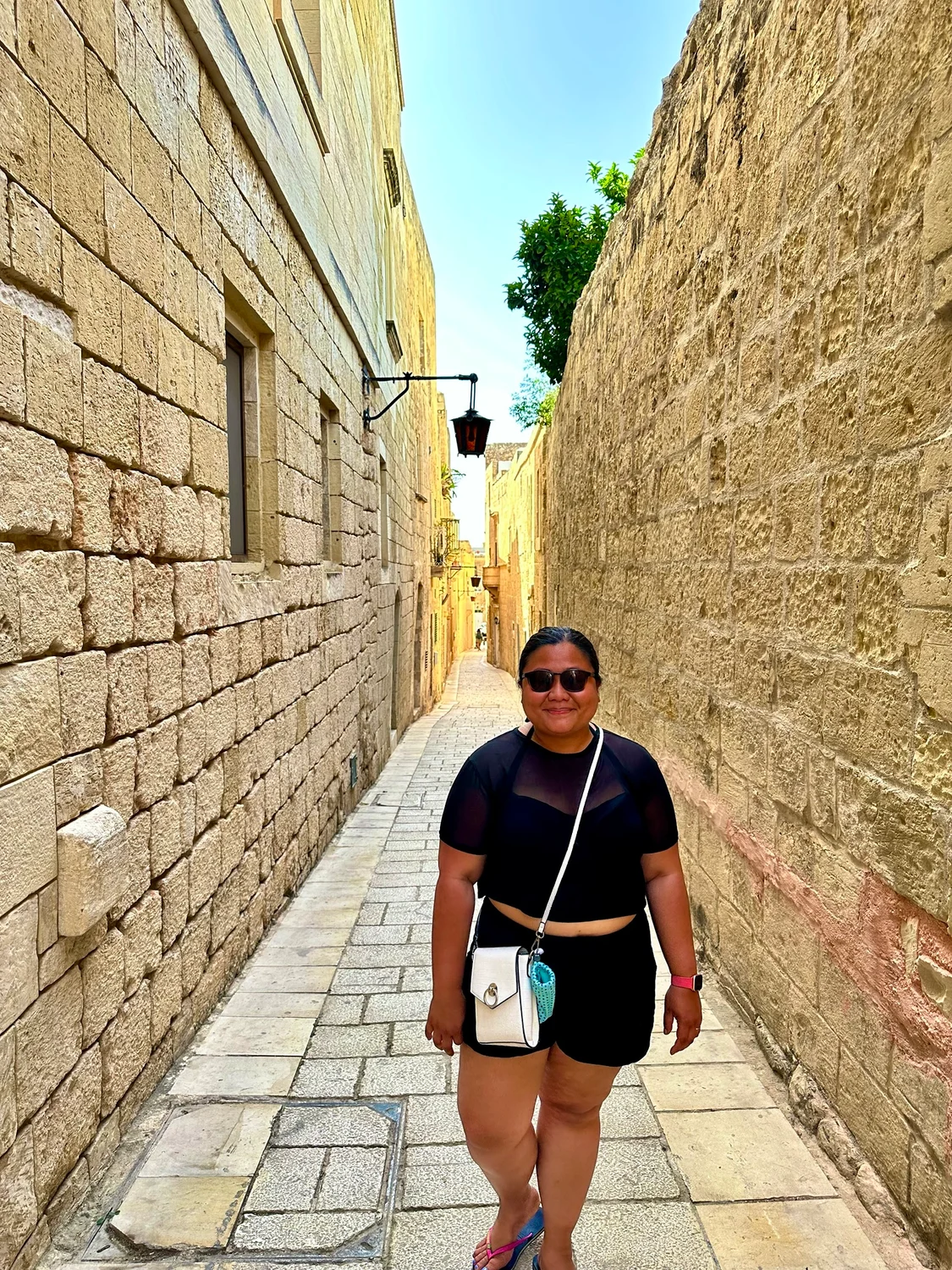 A woman in black shorts and top walks along a narrow stone-paved alley between tall sandstone walls on a sunny day.