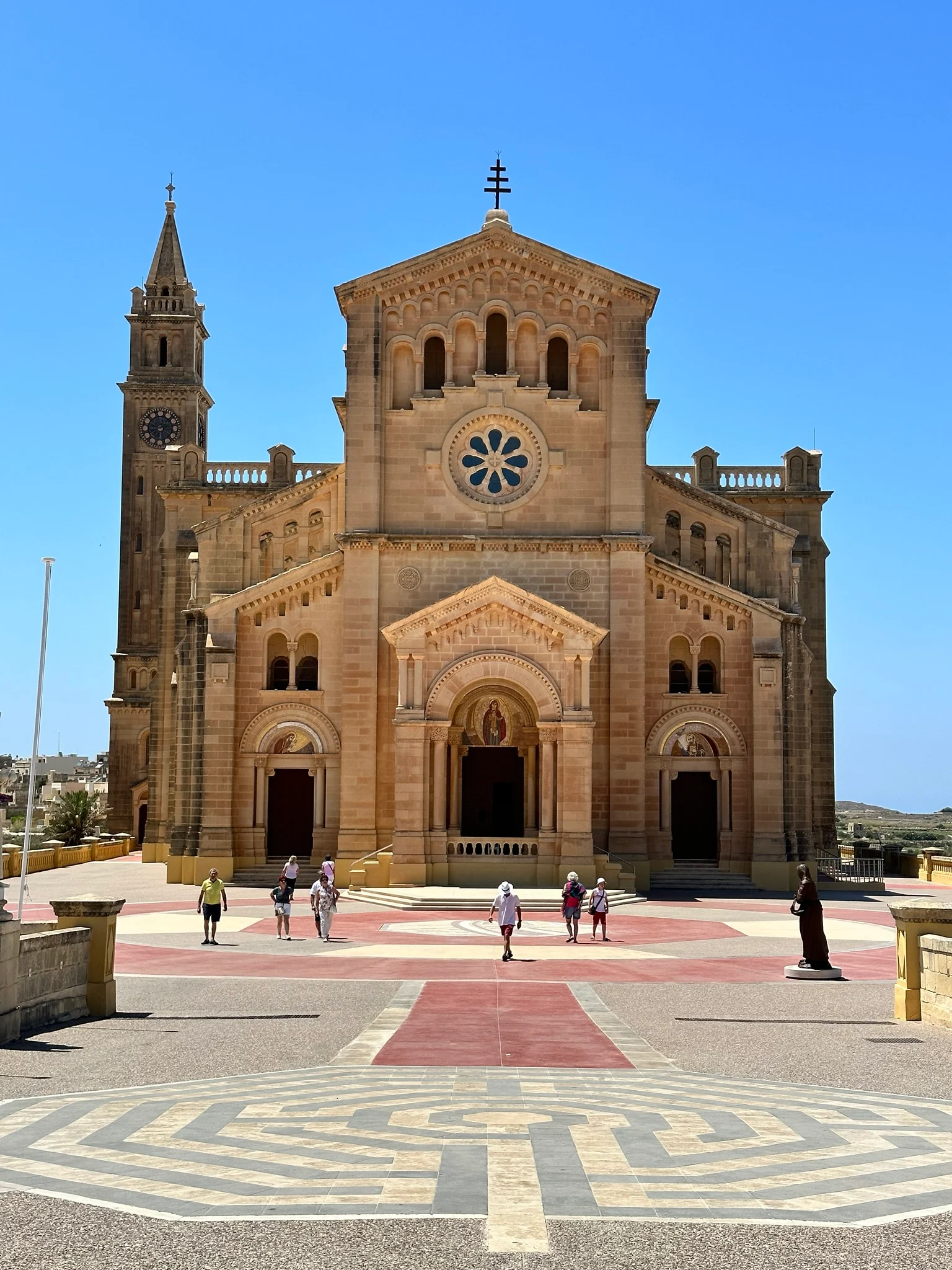 A sandstone basilica with a rose window and bell tower stands under a clear blue sky with visitors crossing the forecourt.