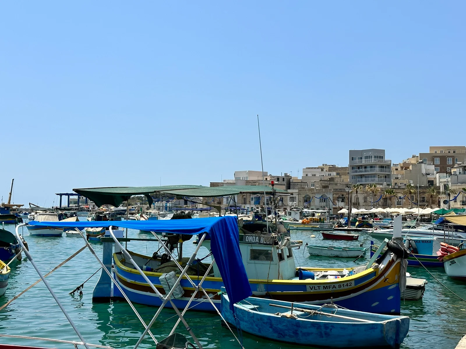 Colourful fishing boats moored in a busy harbour with white stone buildings lining the waterfront under a clear blue sky.