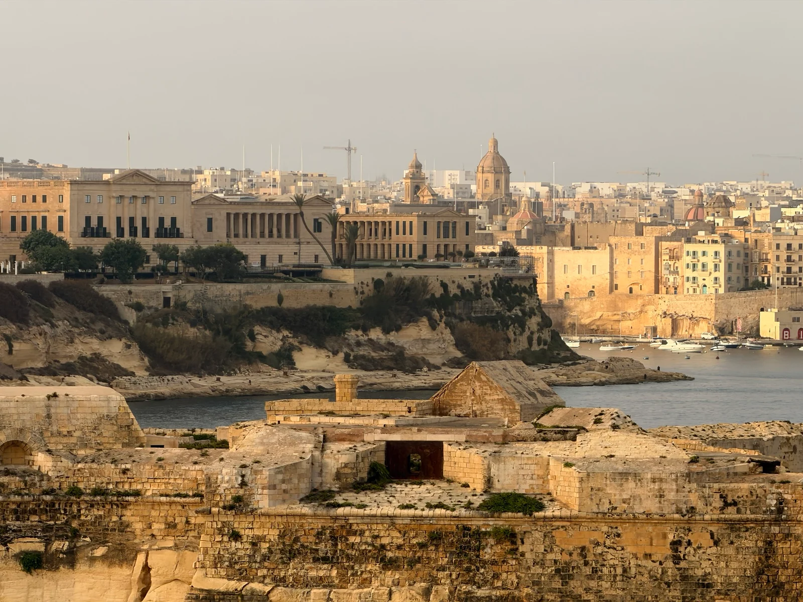 Ancient stone fortifications overlook a calm harbour with a city of pale golden buildings and domed churches beyond.