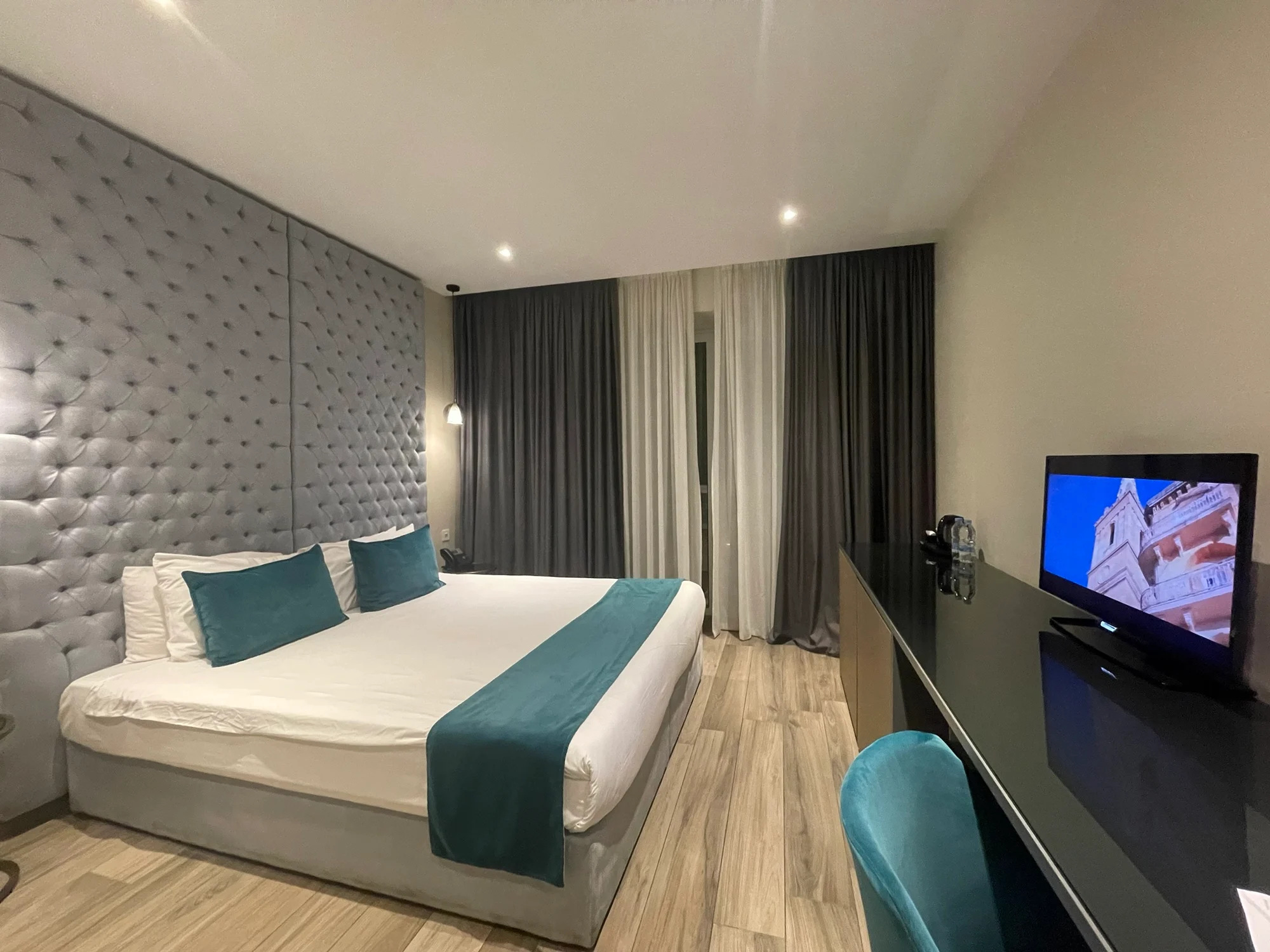 A modern hotel bedroom with a tufted grey headboard, white bedding with teal accents and a flatscreen TV on a dark unit.