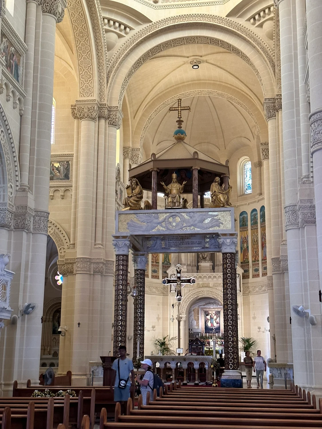 Interior of a basilica with ornate stone arches, gilded statues above the altar and wooden pews in the foreground.