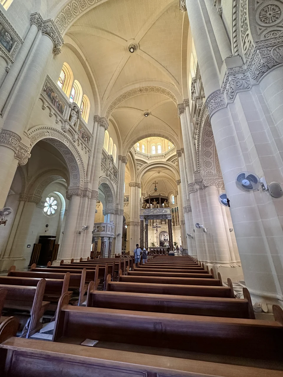 The vaulted stone interior of a basilica with rows of wooden pews leading to an ornate altar beneath a domed ceiling.