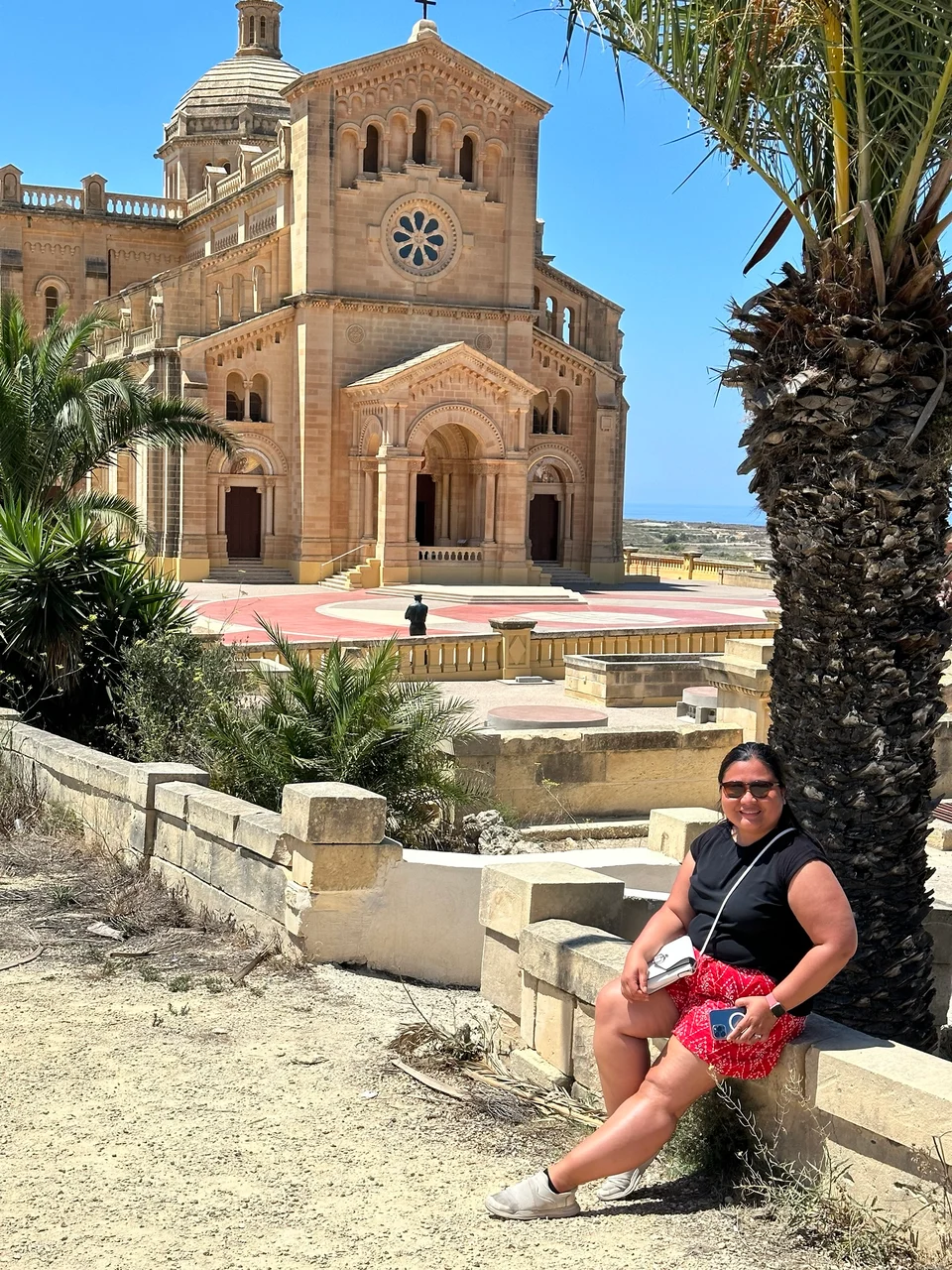 A woman in a black top and red skirt sits on a stone wall beside a palm tree in front of a large honey-coloured basilica.