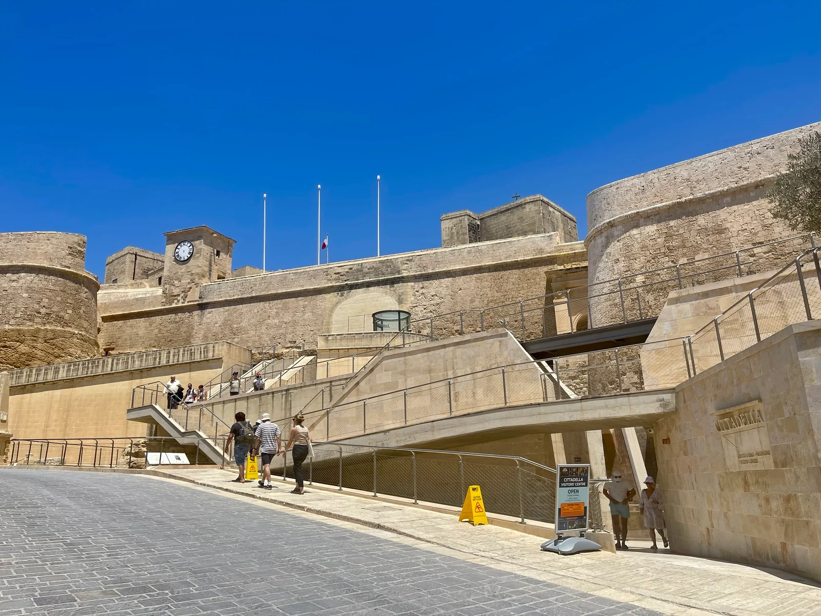 Tourists walk up steps towards the stone walls and towers of the Citadel under a clear blue sky.