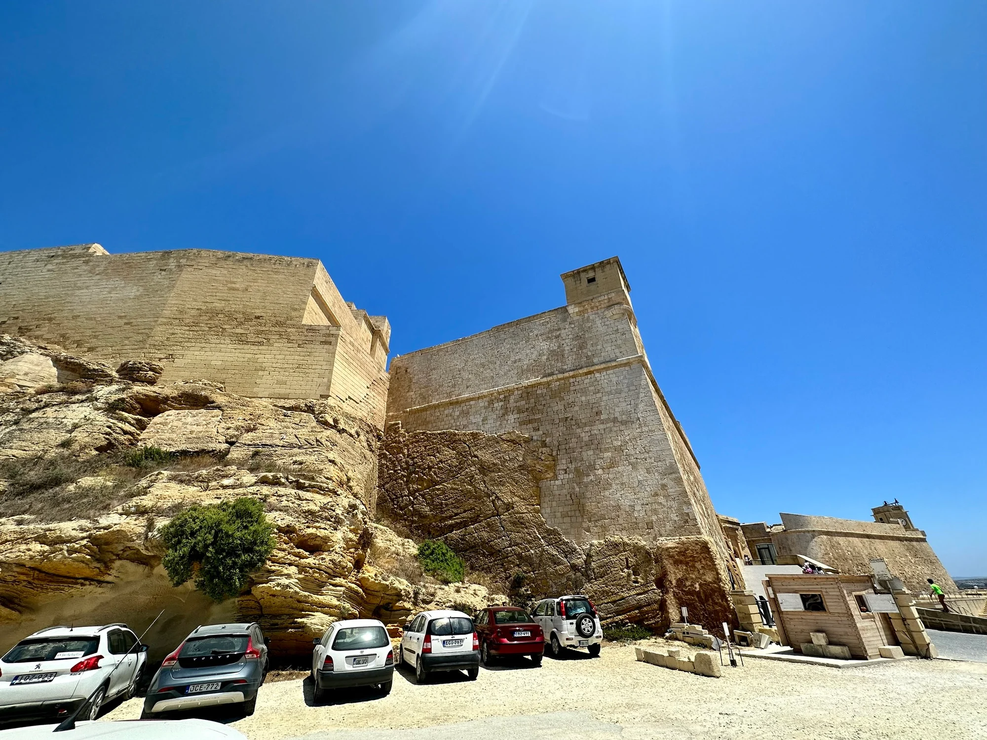 Stone fortress walls rise above rocky outcrops beside a gravel car park with several parked cars under a clear blue sky.