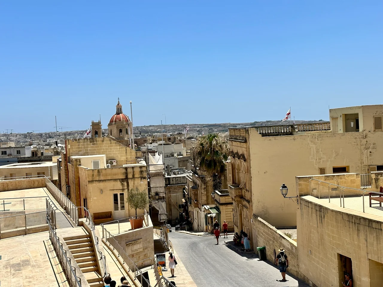 Sunlit limestone buildings and a domed church tower seen from an elevated walkway under a clear blue sky.