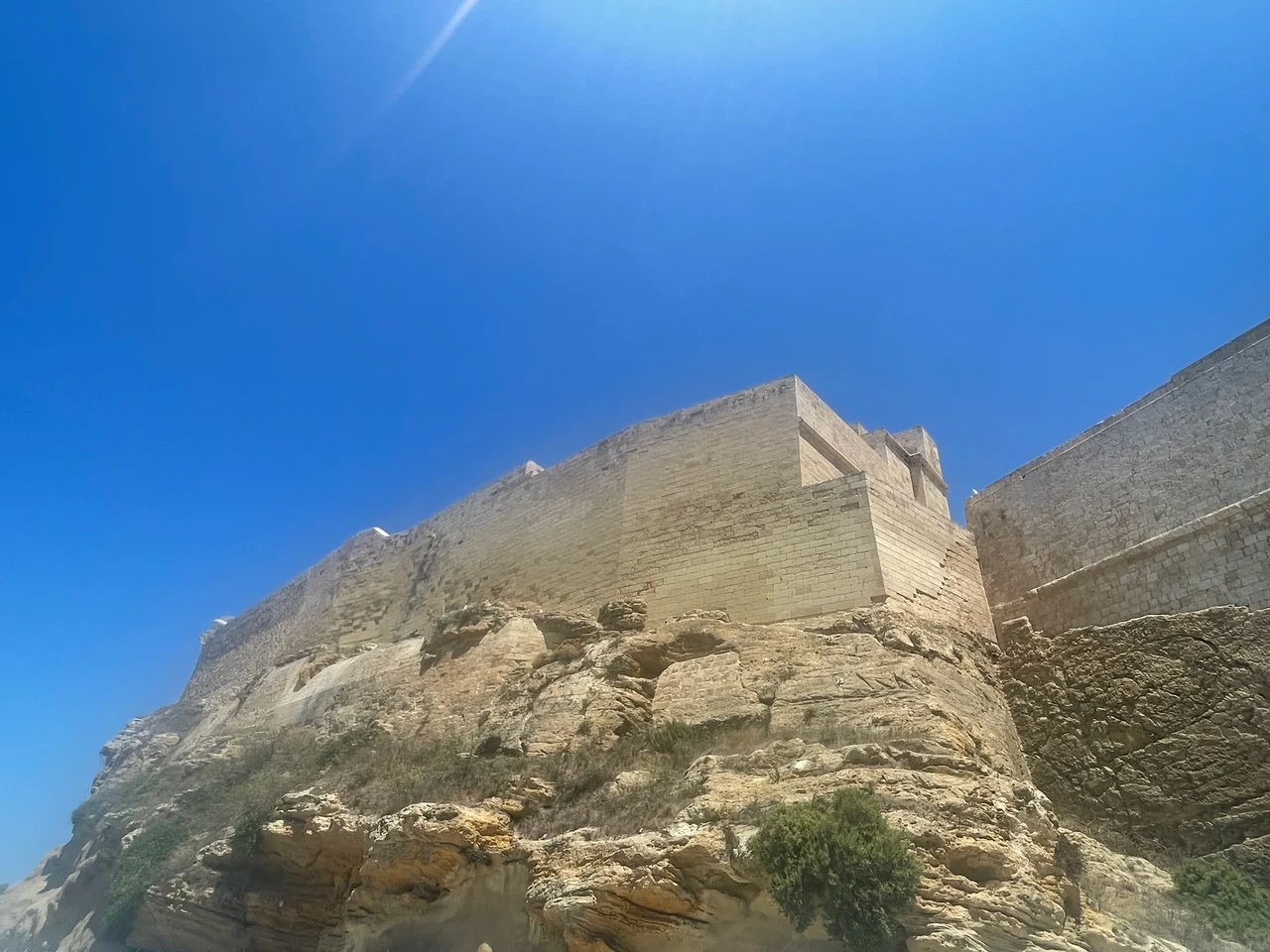 A large ancient stone citadel rises steeply above rough rocky outcrops beneath a clear deep blue sky.
