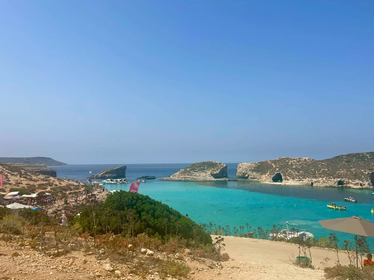 A turquoise lagoon enclosed by rocky limestone headlands under a clear blue sky, with boats and swimmers visible near a sandy shore.