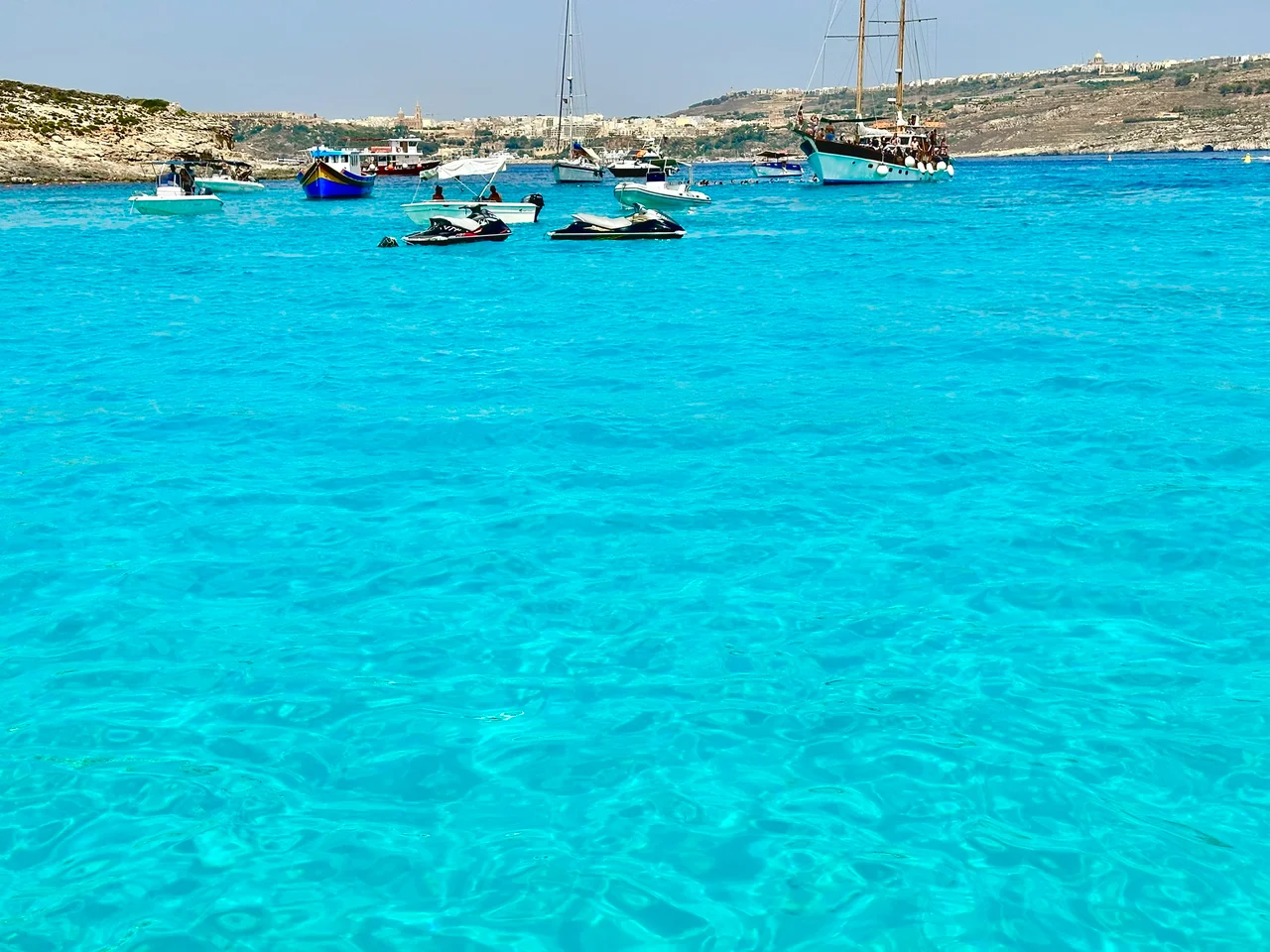 Vivid turquoise water dotted with small boats, jet skis and a tall sailing vessel, with rocky limestone cliffs and a hilltop town beyond.