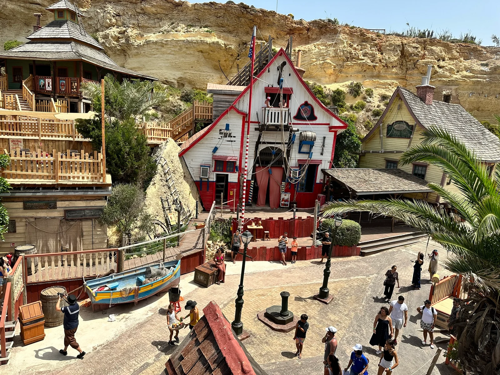 Tourists walk around a quirky seaside village film set with colourful wooden buildings built against a sandy cliff face.