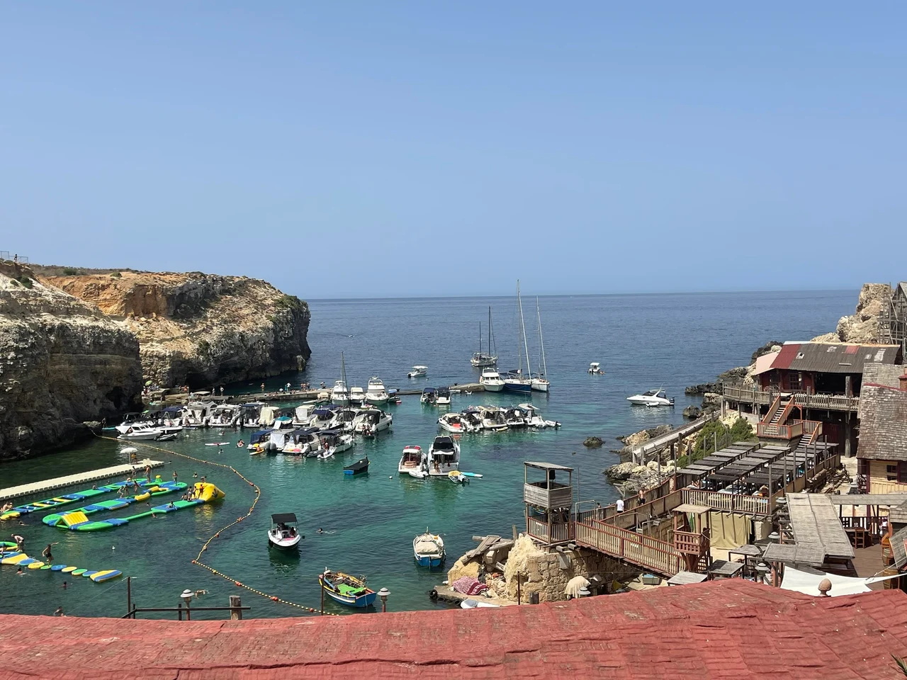 A sheltered cove with turquoise water filled with moored boats, flanked by limestone cliffs and rustic wooden buildings on a sunny day.