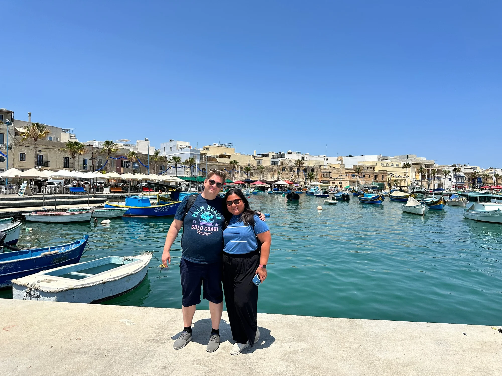 Two people stand arm in arm on a harbour quayside with colourful boats and limestone buildings behind them.