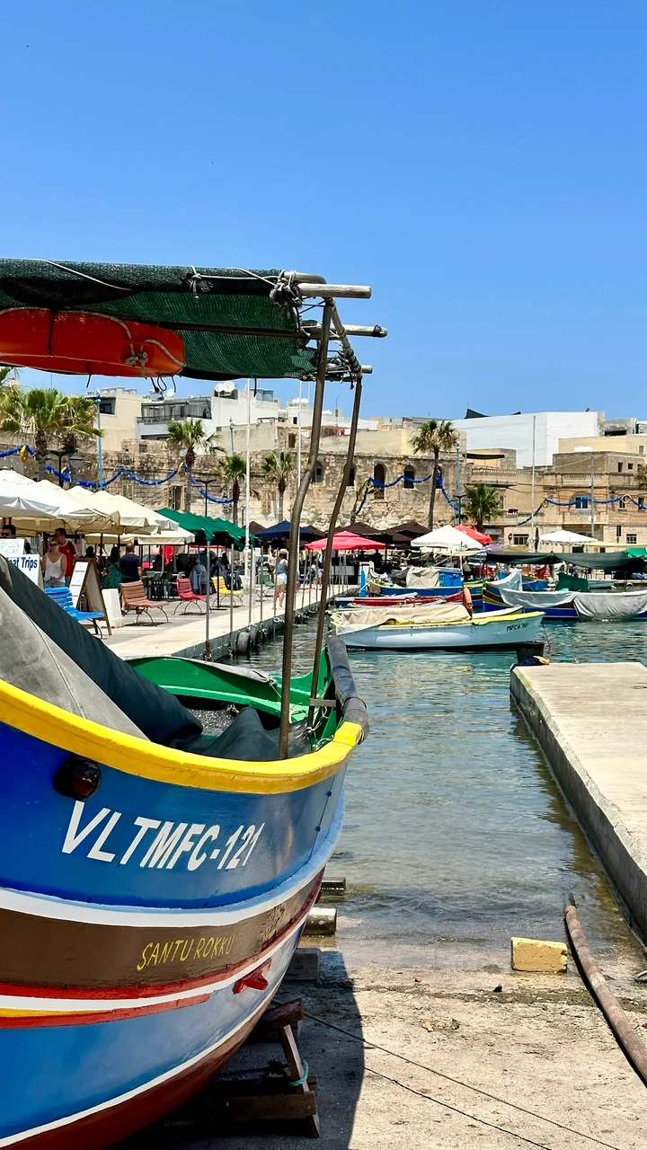 A blue wooden fishing boat named Santu Rokku moored at a sunny harbour with other boats, waterfront cafes and stone buildings beyond.