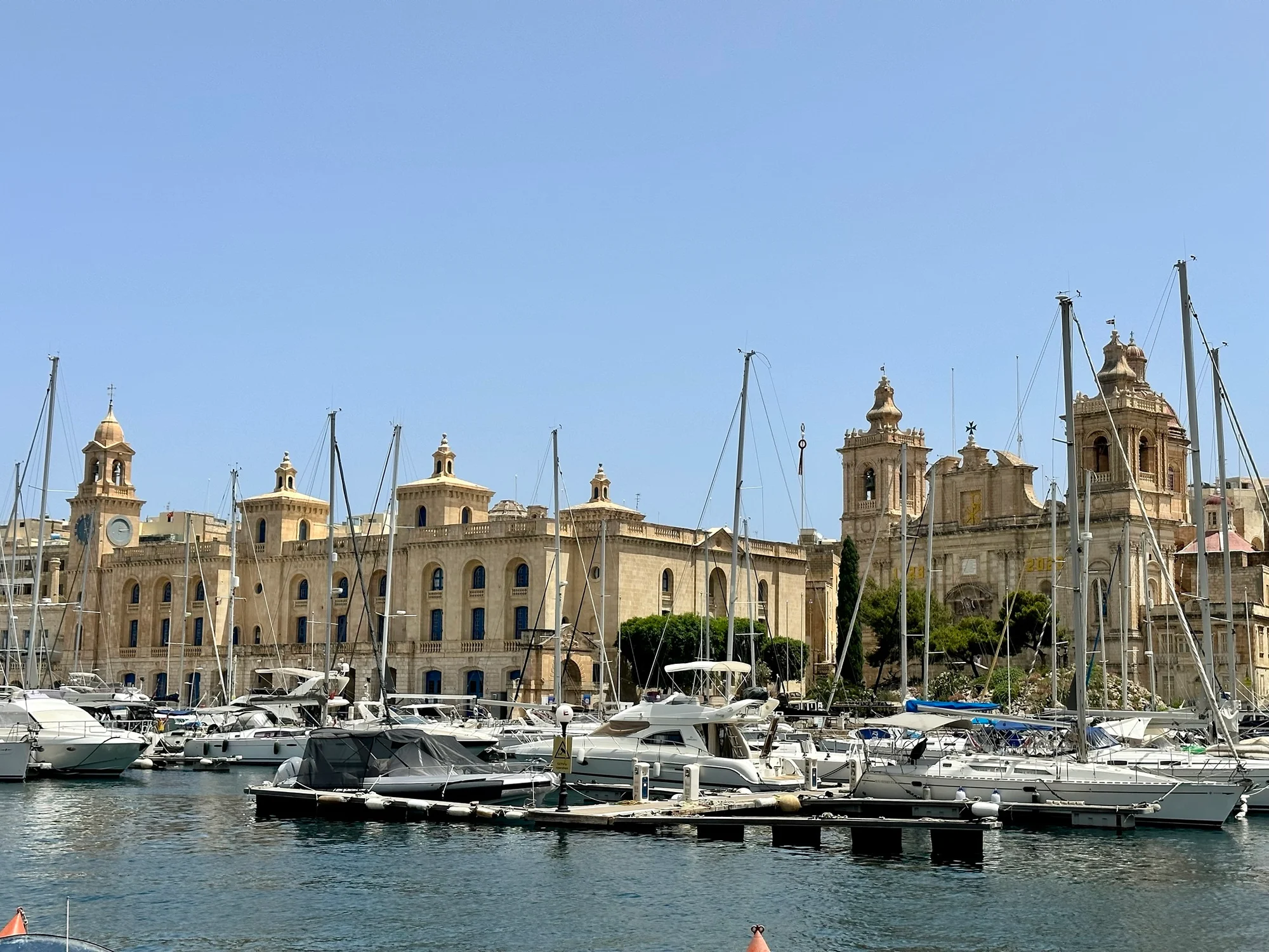 Sailboats moored in a marina in front of large honey-coloured stone buildings with clock towers under a clear blue sky.