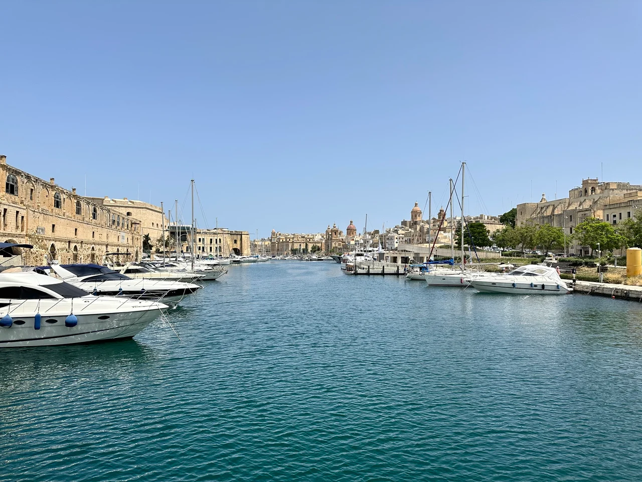 Yachts and motorboats moored along a calm harbour channel flanked by honey-coloured stone buildings under a clear blue sky.