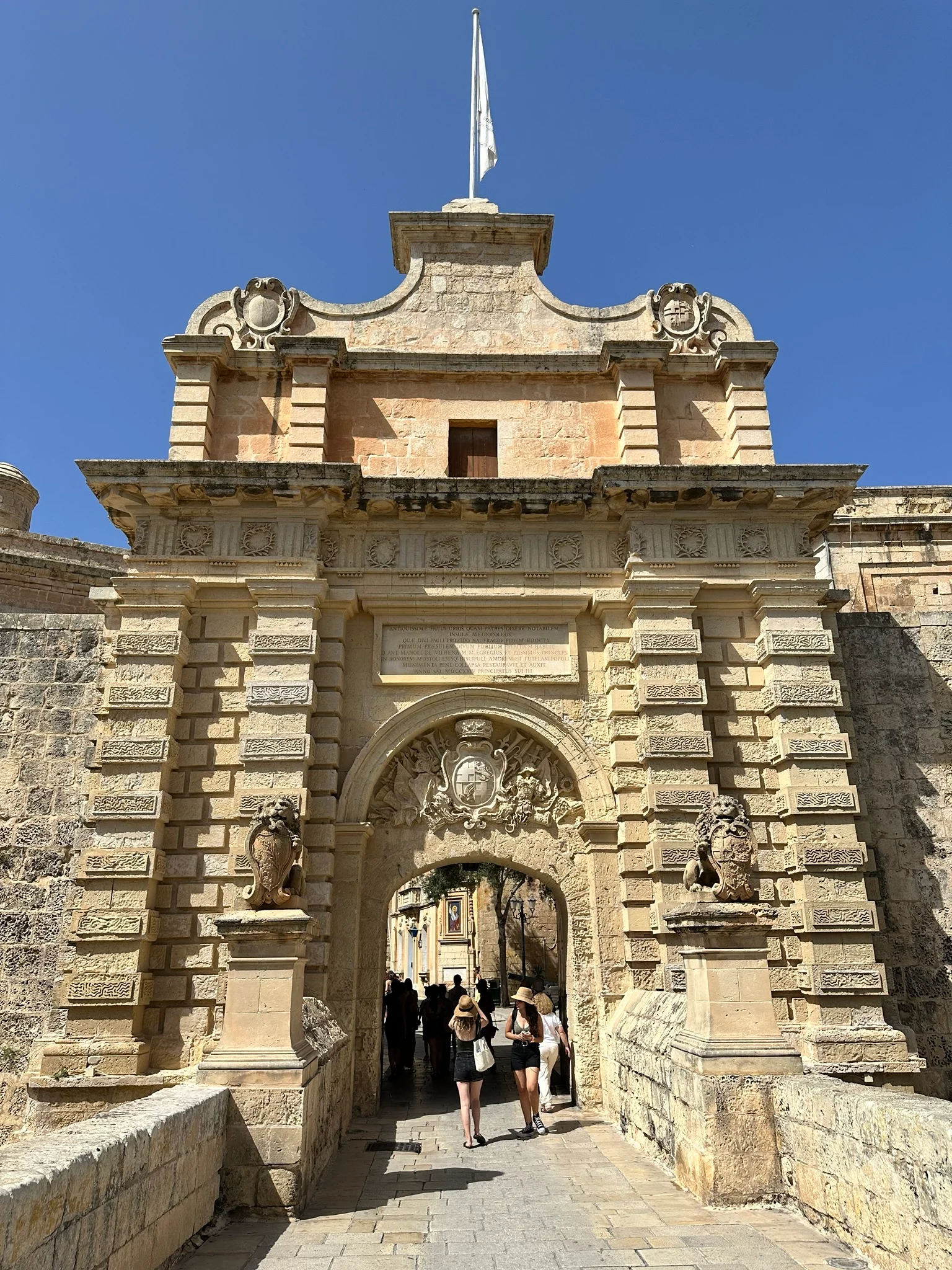 Tourists walking through an ornate stone arched gateway decorated with carved lions and a coat of arms under a clear blue sky.